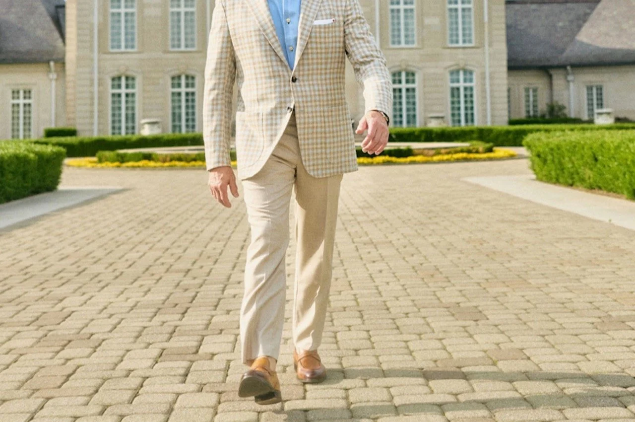 Man walking on cobblestone driveway in front of large mansion, dressed in beige and blue suit.