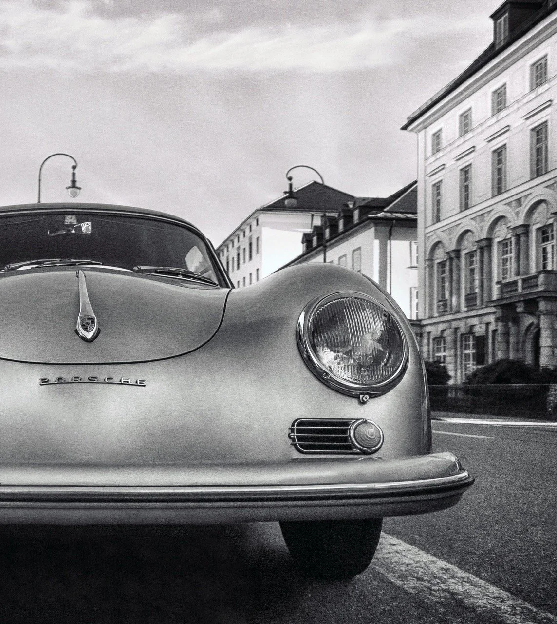 Grayscale photo of a vintage Porsche car parked on a city street with historic buildings in the background.