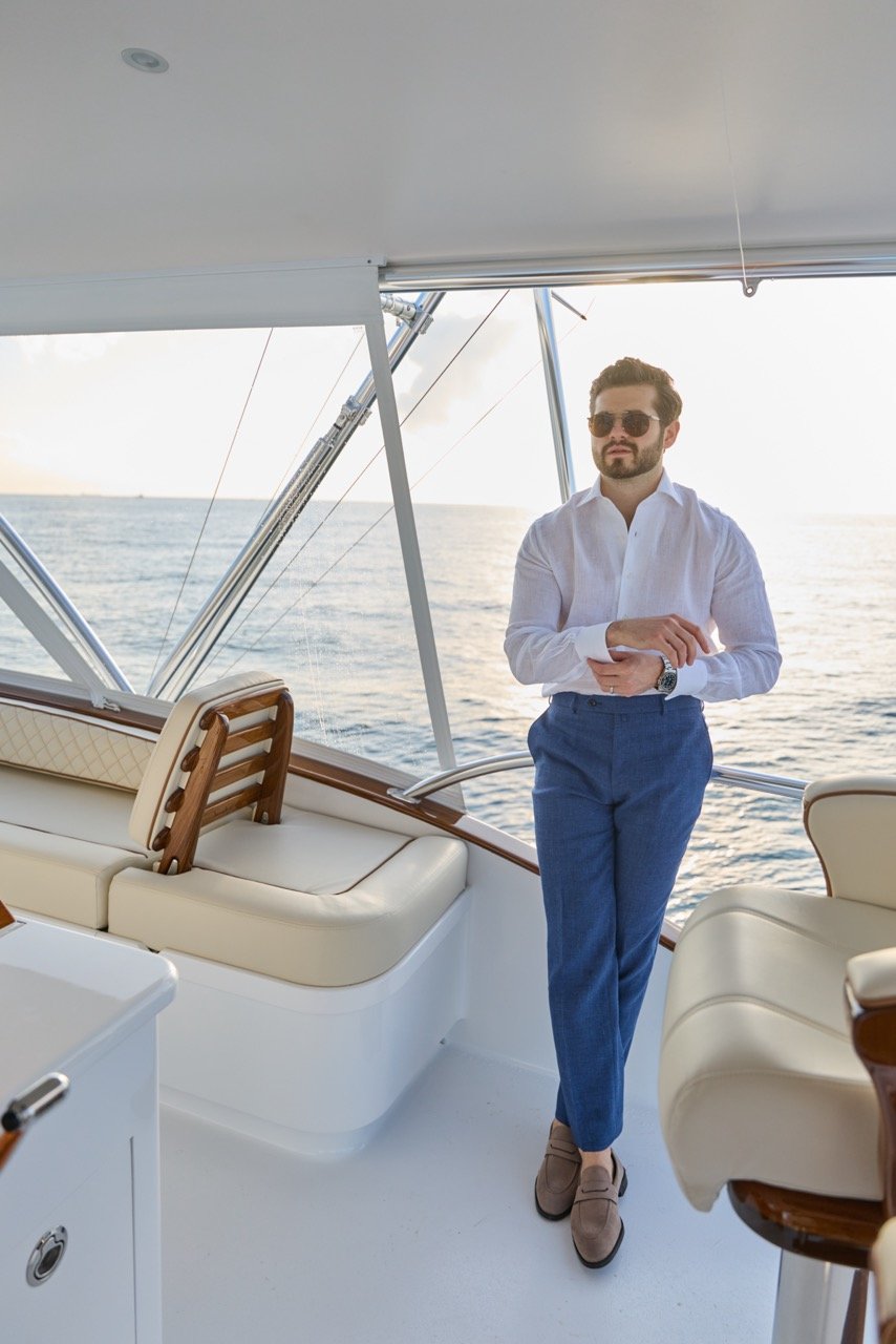A man in a white shirt, blue pants, and sunglasses standing on a yacht with the ocean in the background during sunset.