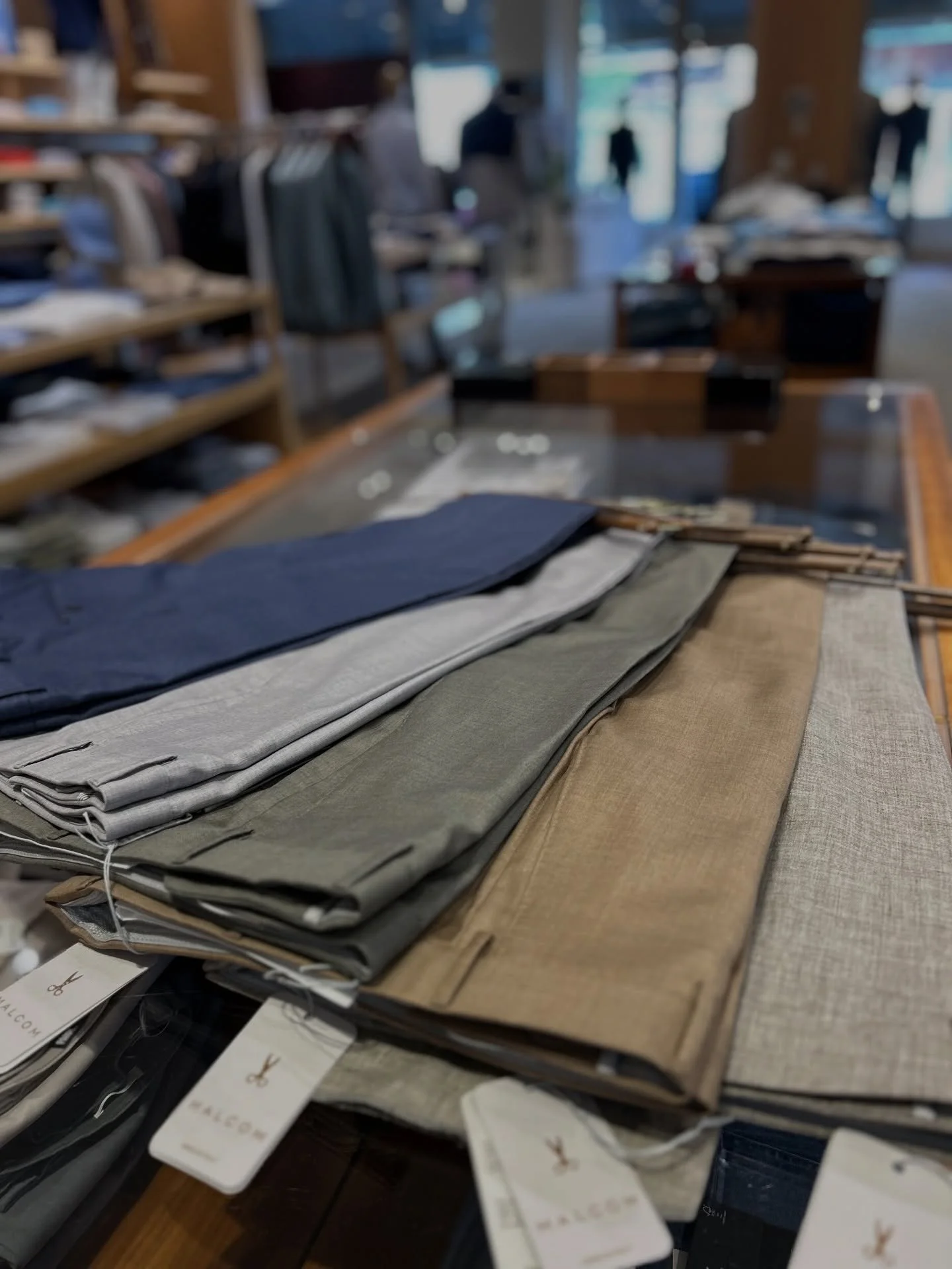 Stacks of folded fabric and clothing samples on a wooden table in a boutique or fabric store, with tags and labels visible.