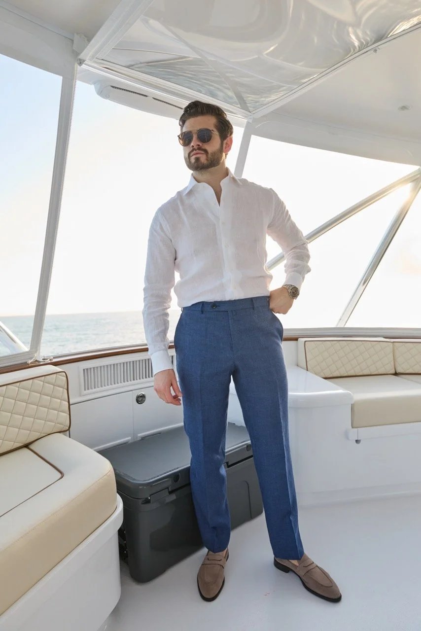 A man with dark hair and sunglasses wearing a white shirt and blue trousers standing on a boat with ocean in the background.