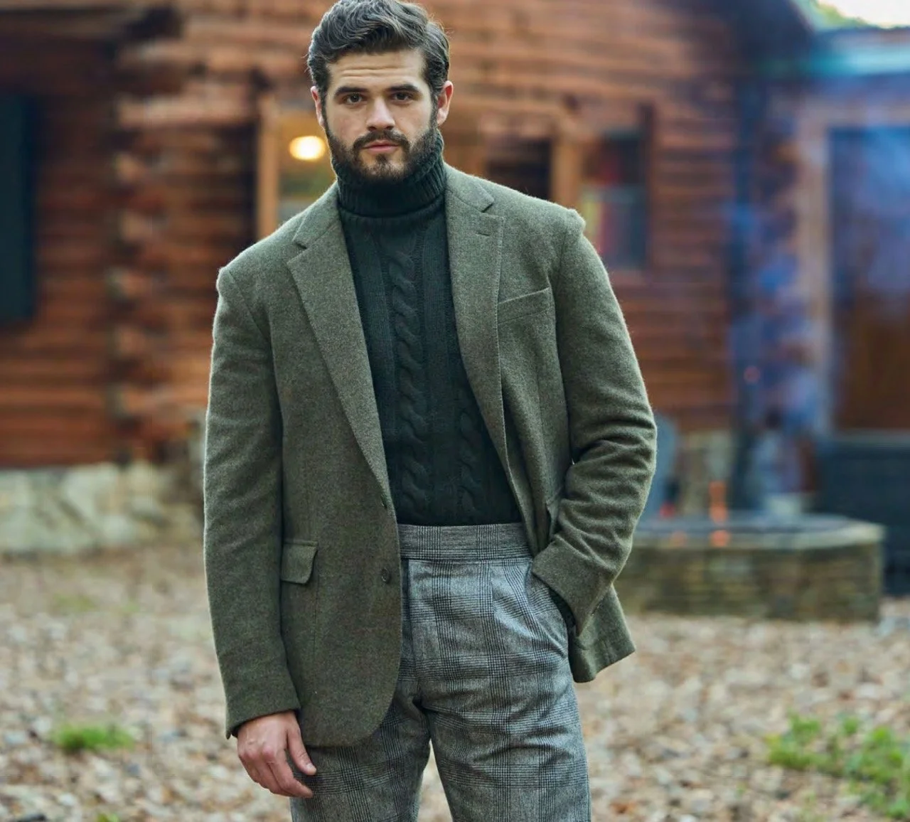 A man with dark hair and a beard dressed in a green blazer, black turtleneck sweater, and gray plaid trousers, standing outdoors in front of a wooden house.