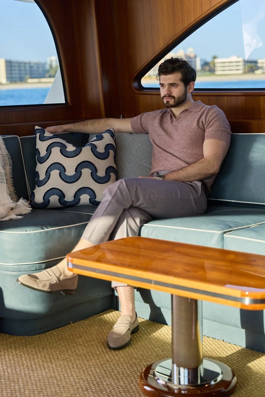 A young man with dark hair and a beard sitting on a turquoise sofa inside a boat, with a view of city buildings and water through the windows. He is wearing a light brown polo shirt, light-colored pants, and beige loafers. His arm is resting on a dec