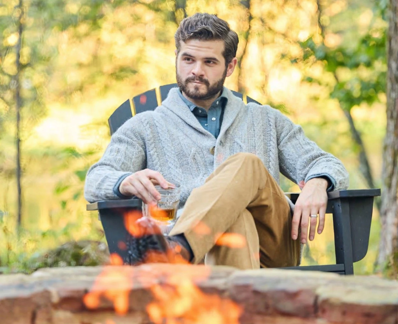 A man with a beard and brown hair sitting in a black chair outdoors during autumn, holding a glass of whiskey, with a campfire in front of him and trees with yellow leaves in the background.