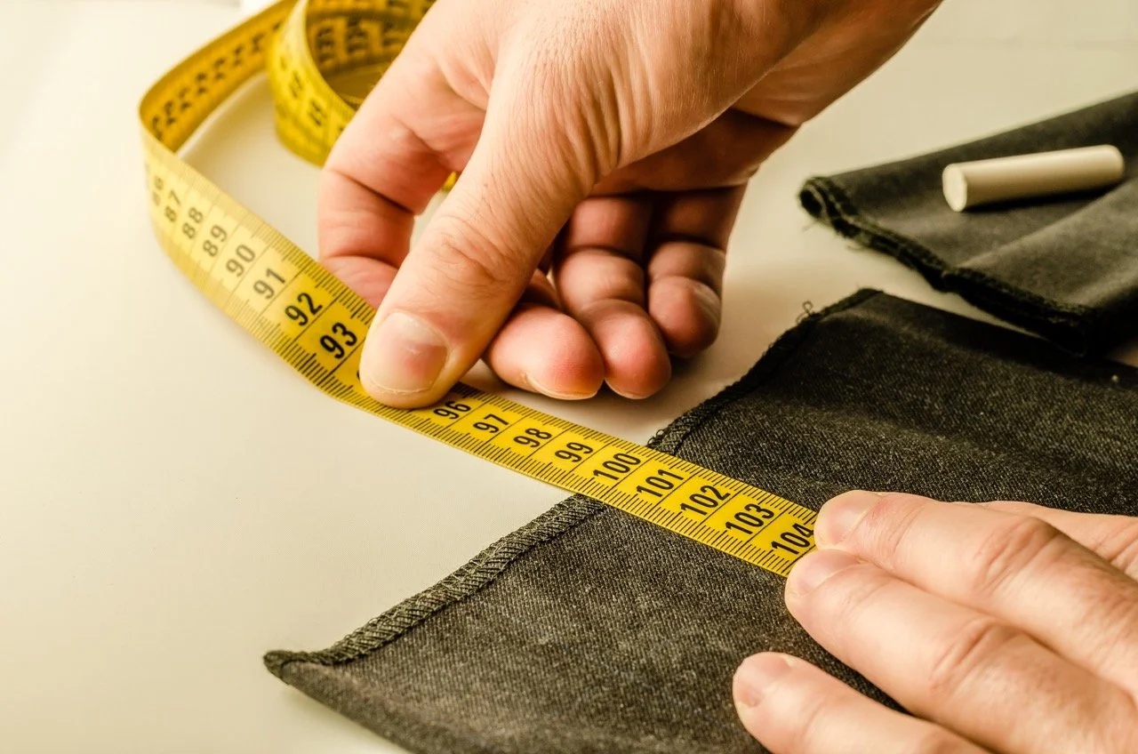 Hands measuring a piece of dark fabric with a yellow tape measure on a white surface.