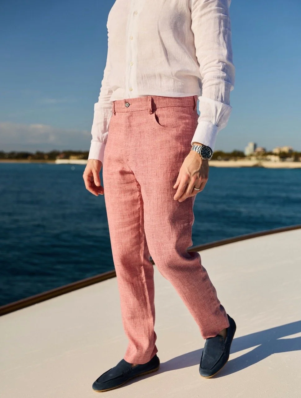 Person standing on a boat near the water, wearing pink pants, a white shirt, and loafers, with a watch on their left wrist.