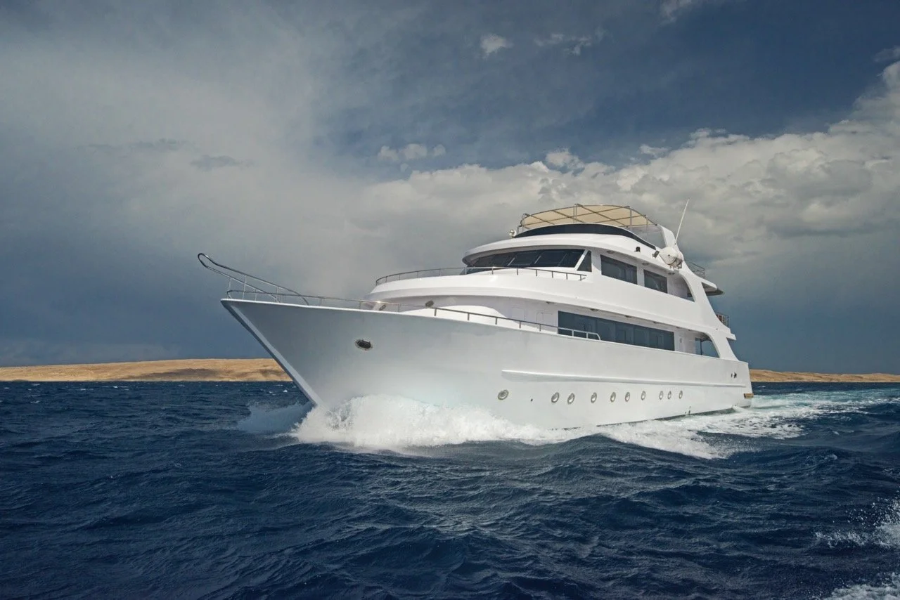 A large white yacht sailing on the ocean with a cloudy sky and a shoreline in the background.