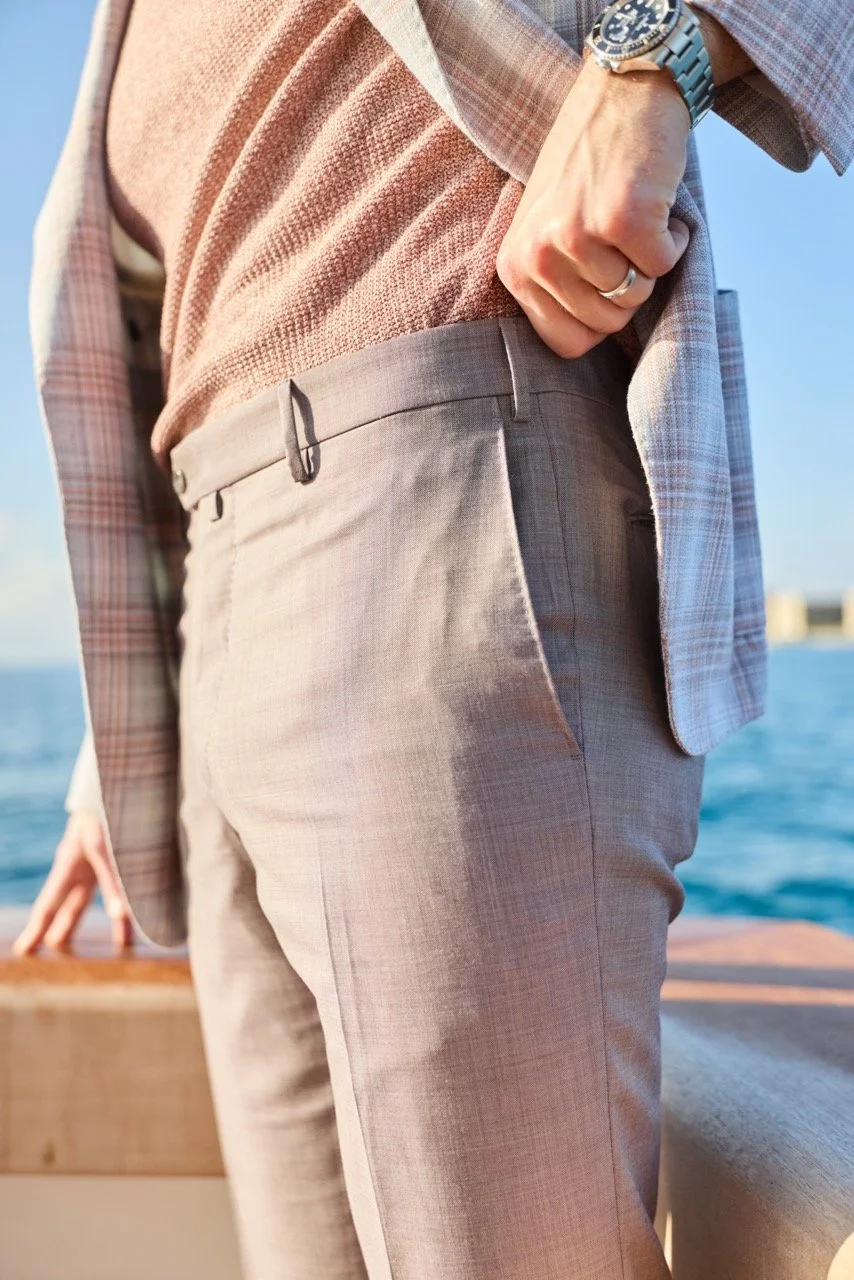 Close-up of a man's styled outfit with beige trousers, a pink patterned sweater, and a plaid blazer, standing outdoors by the water during sunset, showing his hand with a silver watch and wedding ring.