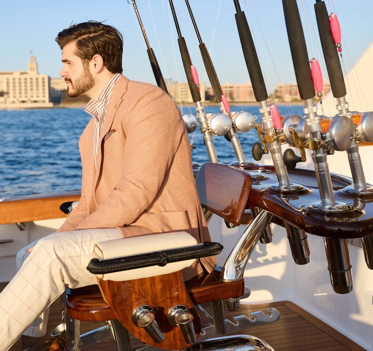 A man with brown hair, a beard, and dressed in a tan blazer and light checkered pants sitting on a boat near fishing rods, with water and city buildings in the background.