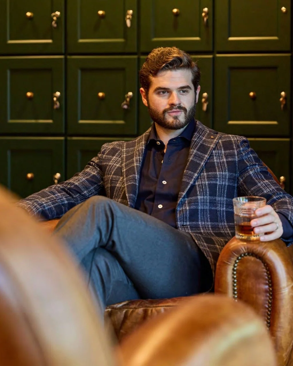A man with dark hair and a beard sitting on a leather chair in a room with green cabinets in the background, holding a glass of whiskey.
