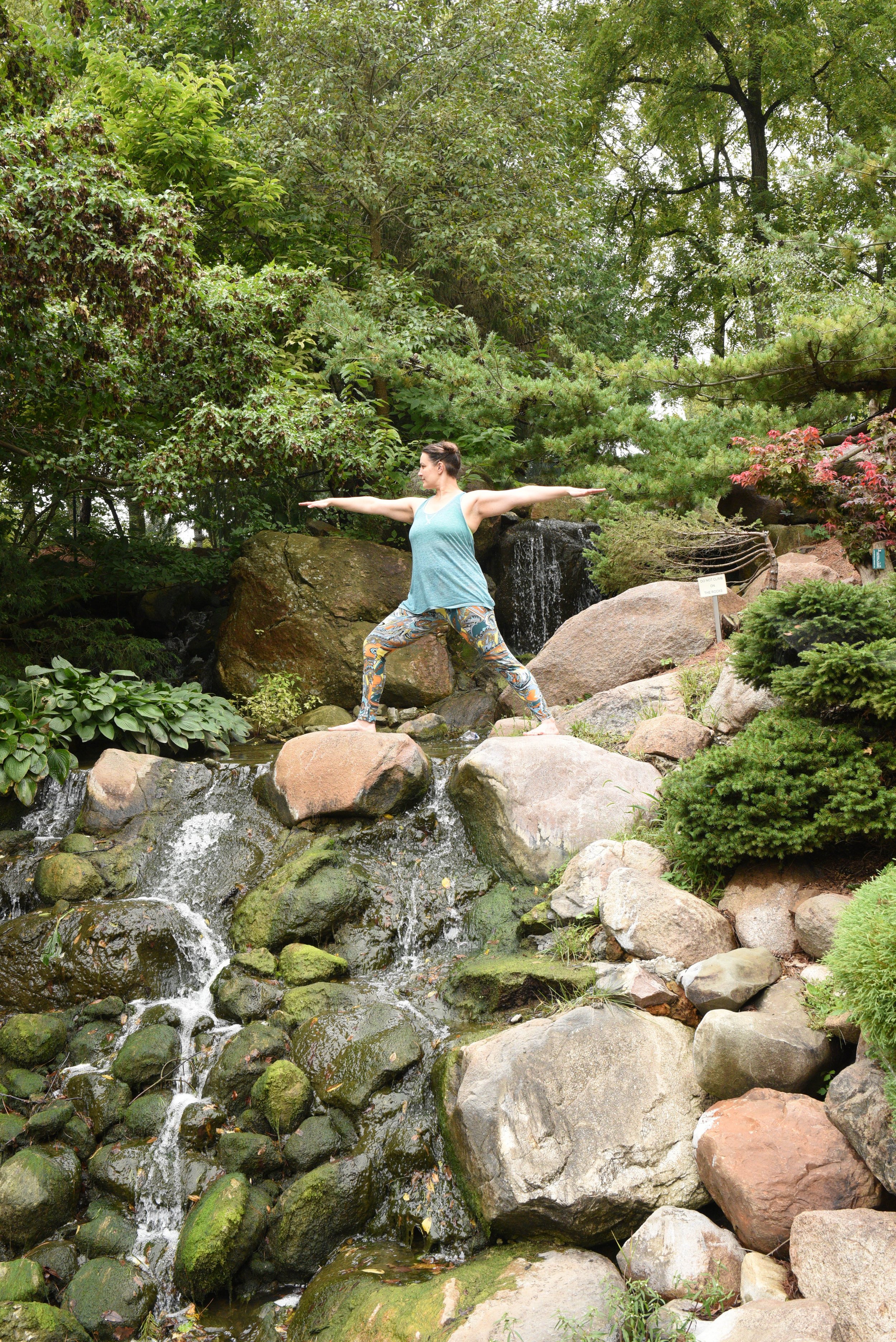 A woman practicing yoga in a tree pose on rocks next to a small waterfall surrounded by lush green plants and trees.