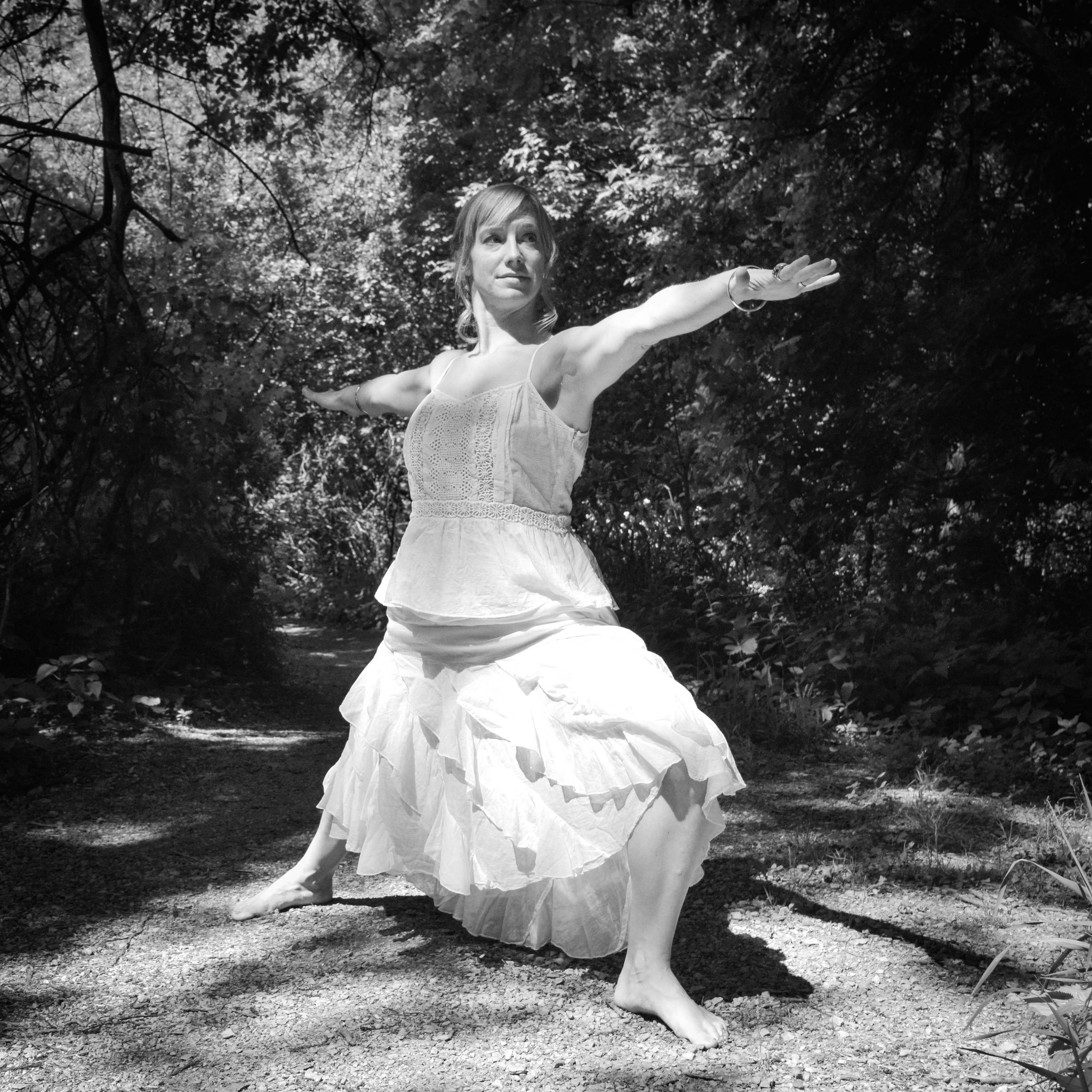 A woman in a flowing dress practicing martial arts or a dance pose in a wooded outdoor setting