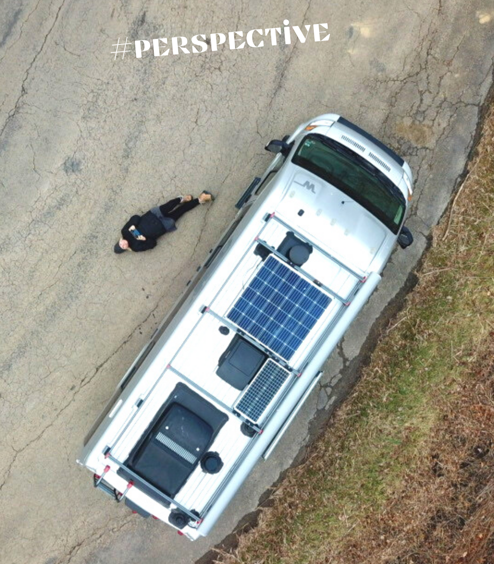 An aerial view of a person lying on the cracked pavement beside a sprinter van equipped with solar panels on its roof.