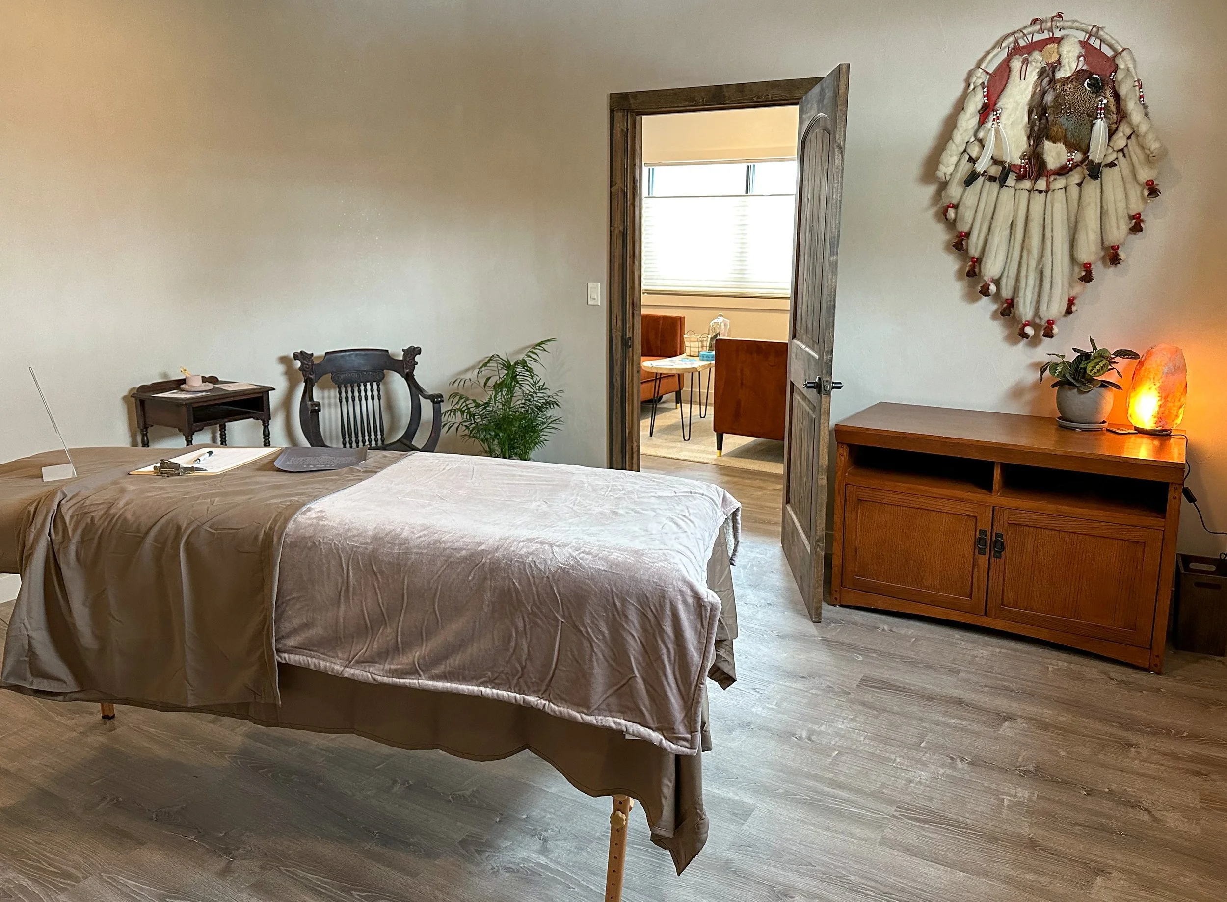 Room with a bed, a wooden cabinet with a plant and salt lamp, wall hanging, open door to a sitting area with table and chairs, hardwood floor, minimal decor.