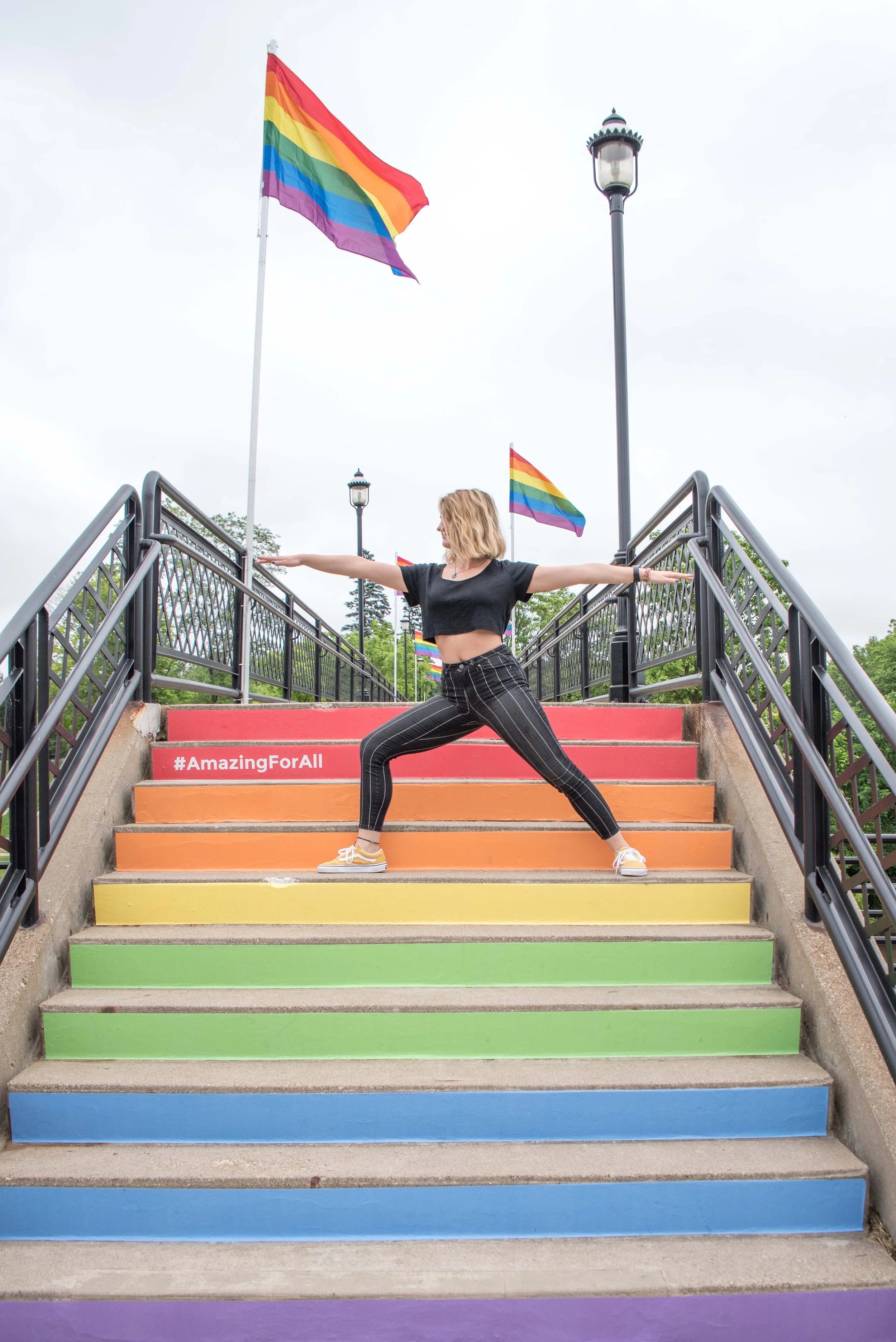 Woman in black clothing performing yoga pose on rainbow-colored stairs at pride event with rainbow flags and lampposts in background.