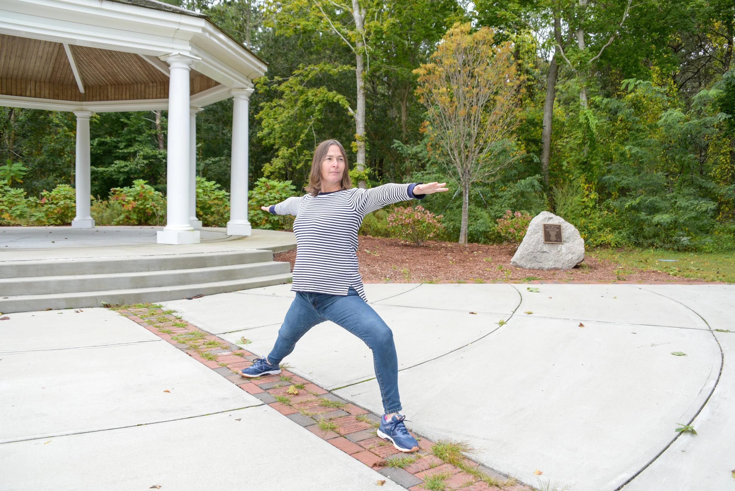 Woman practicing Warrior II yoga pose outdoors in front of a white gazebo with trees in the background.