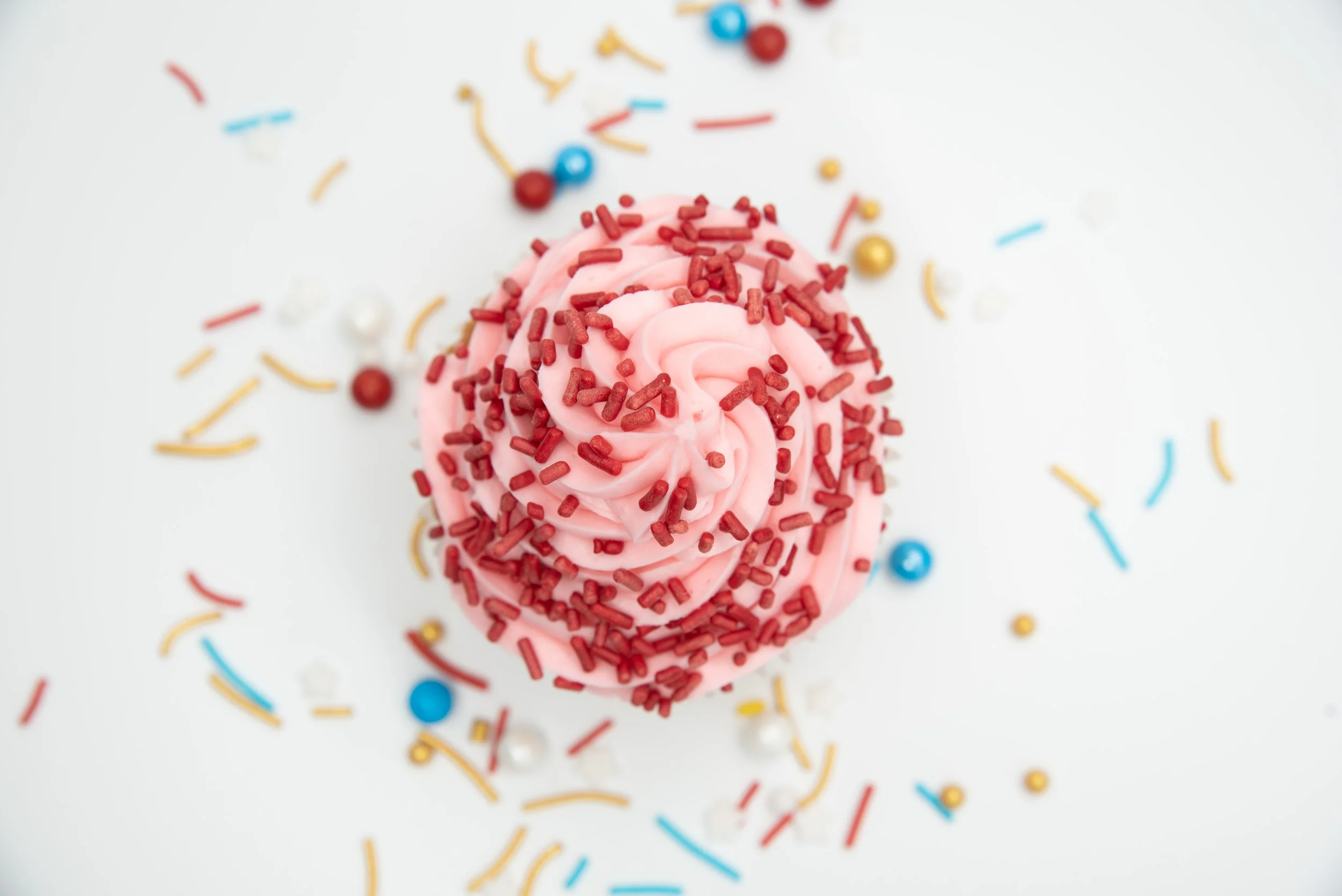 Pink cupcake with red sprinkles surrounded by colorful confetti and small candies on a white background.