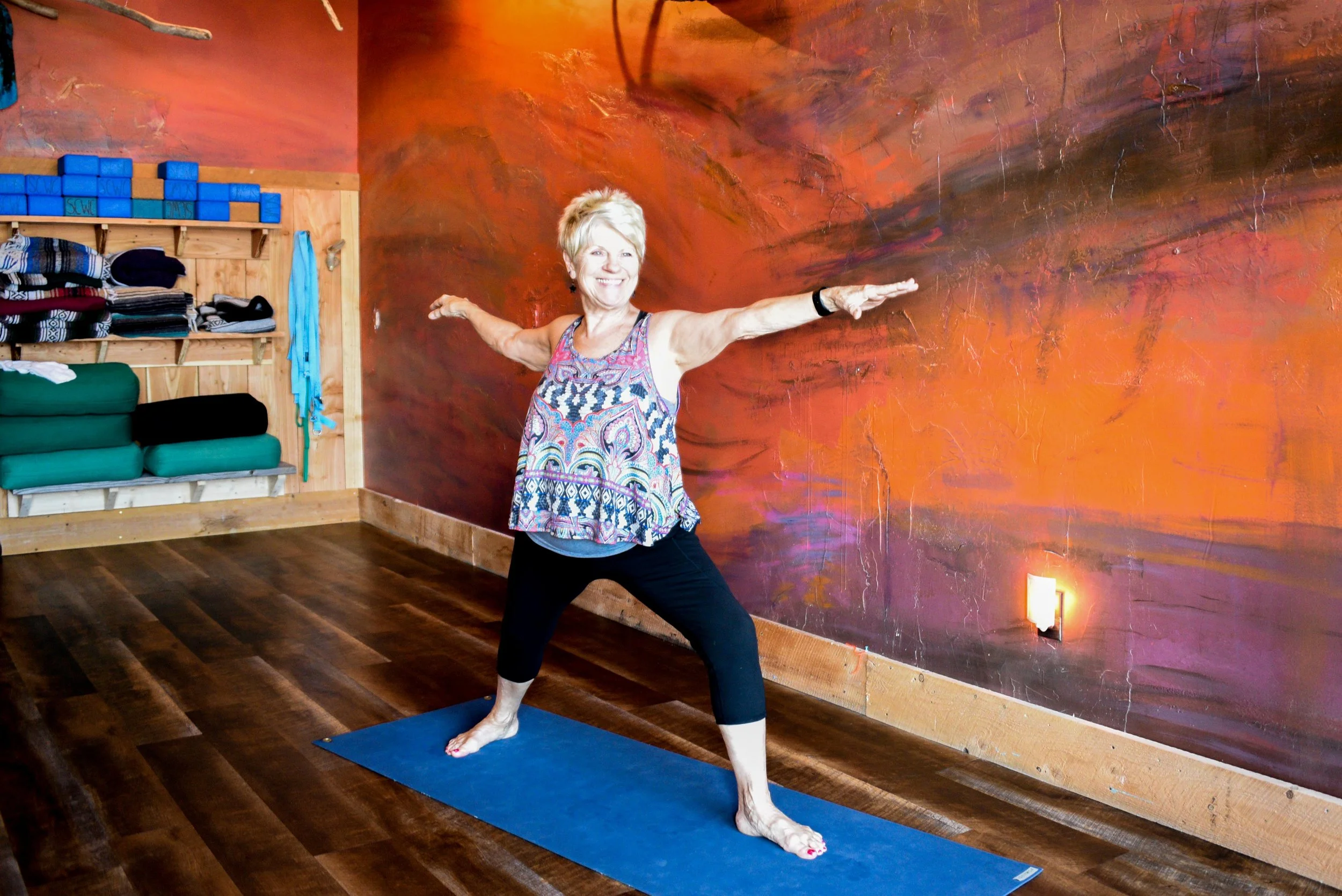A woman practicing Warrior II yoga pose in a studio, standing on a blue yoga mat with arms extended outward, smiling, and wearing a colorful, patterned tank top and black leggings.