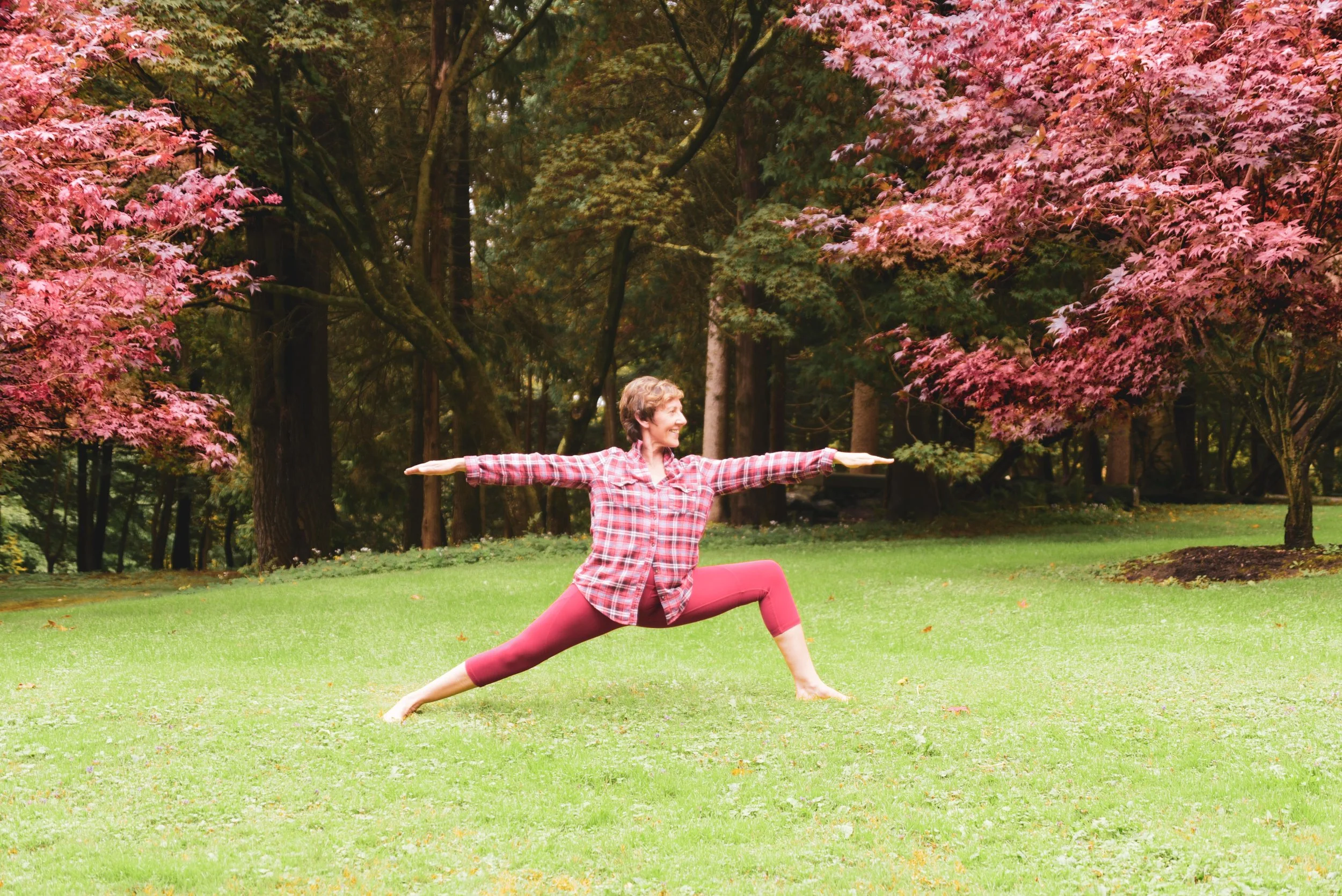 A woman practicing Warrior II yoga pose outdoors on a grassy field surrounded by trees with pink and green leaves.