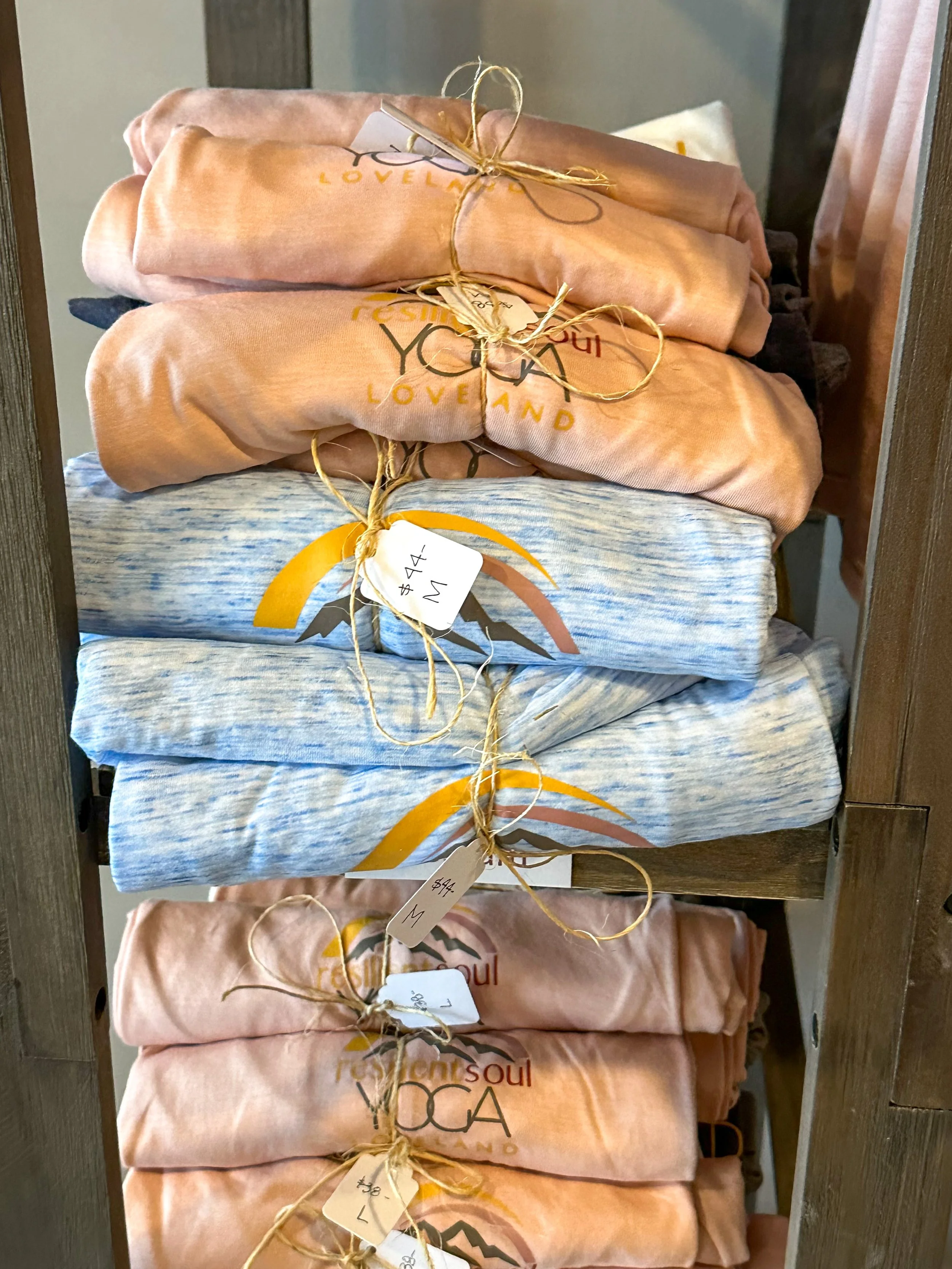 Stacked folded T-shirts or sweatshirts with various patterns and colors, tied with string and sale tags, on a wooden shelf.