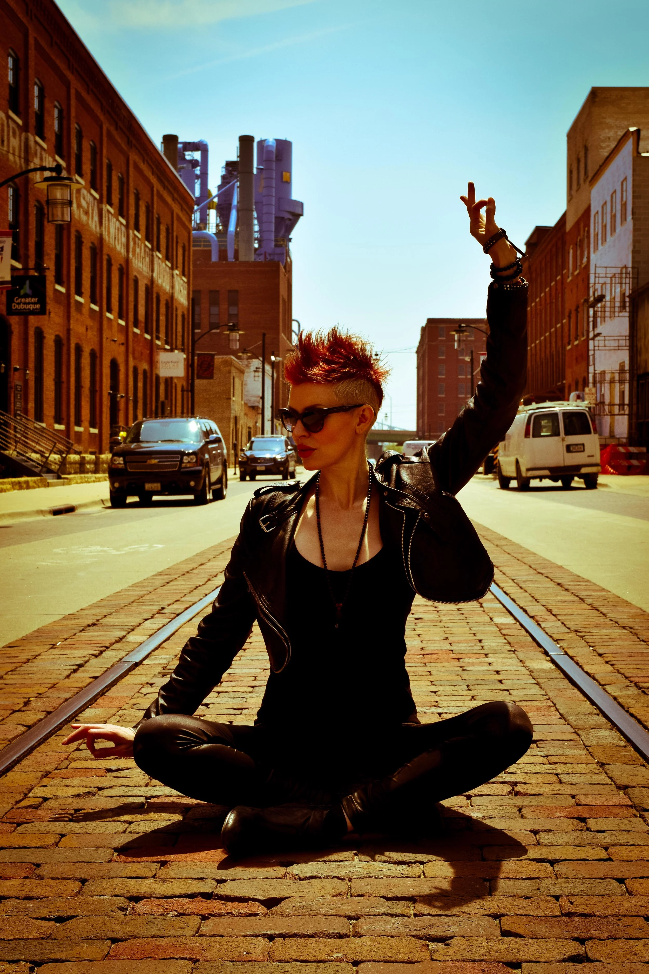 A woman with short red spiky hair, wearing sunglasses and a black leather jacket, sits cross-legged on a cobblestone street with train tracks. She is making a peace sign with her right hand raised in the air and appears to be meditating or posing in an urban setting with cars and brick buildings.