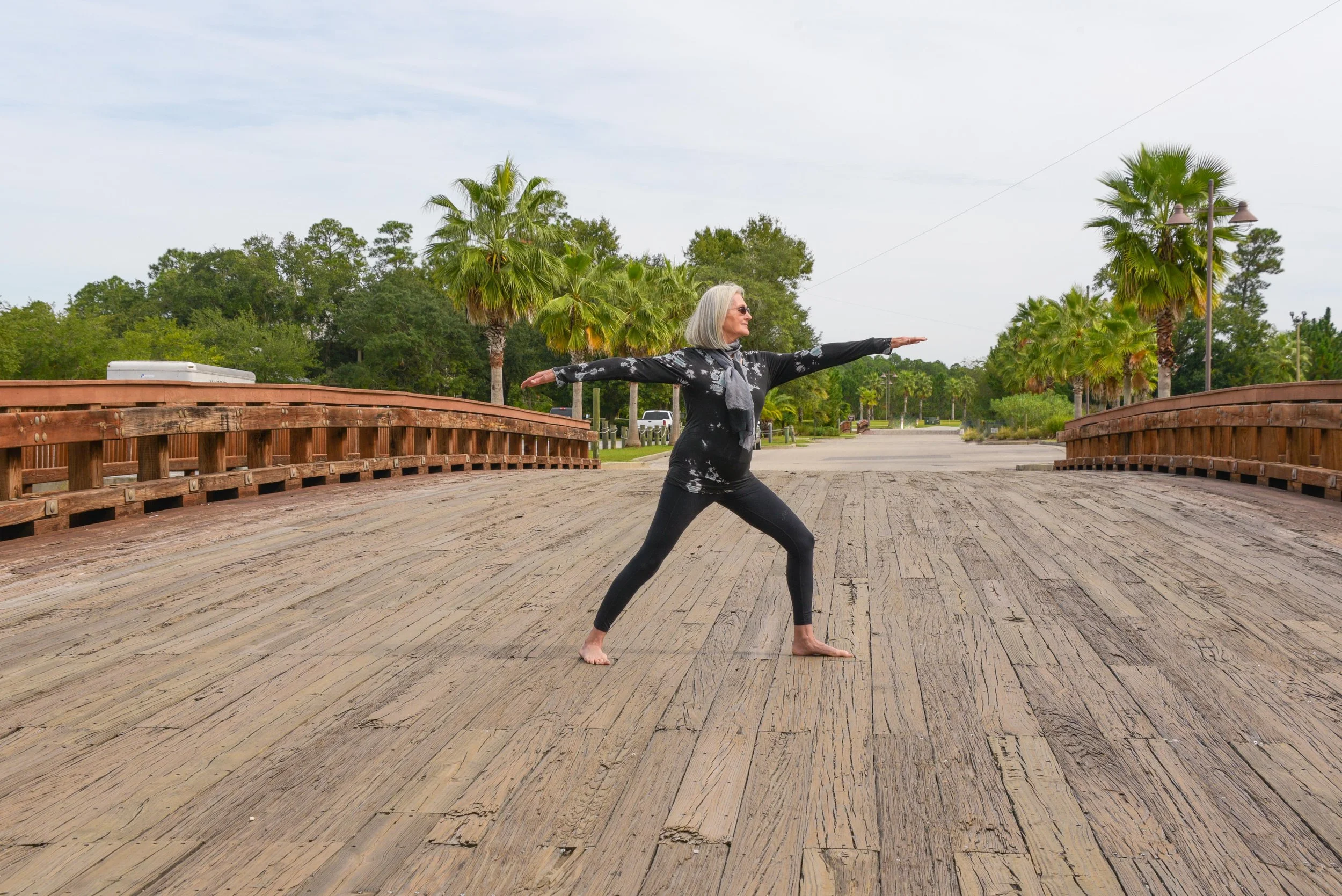 An older woman practicing the warrior II yoga pose on a wooden bridge surrounded by palm trees and lush greenery.