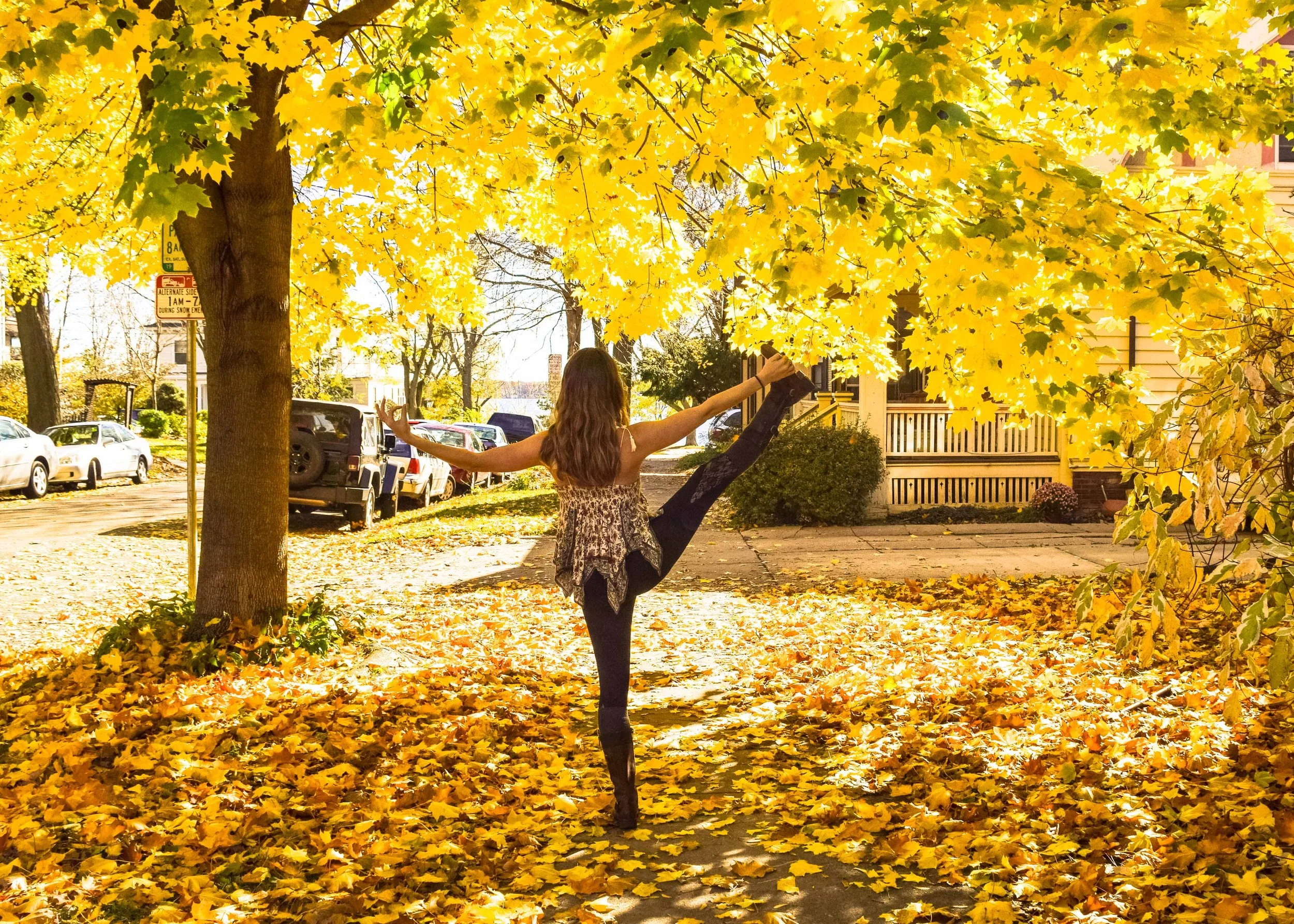 A girl practicing a high kick under a large tree with yellow leaves, on a sidewalk covered with fallen leaves, in a residential neighborhood.