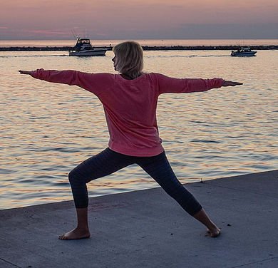 A woman in a red long-sleeve shirt and black leggings practicing yoga near the water at sunset, with boats in the background.