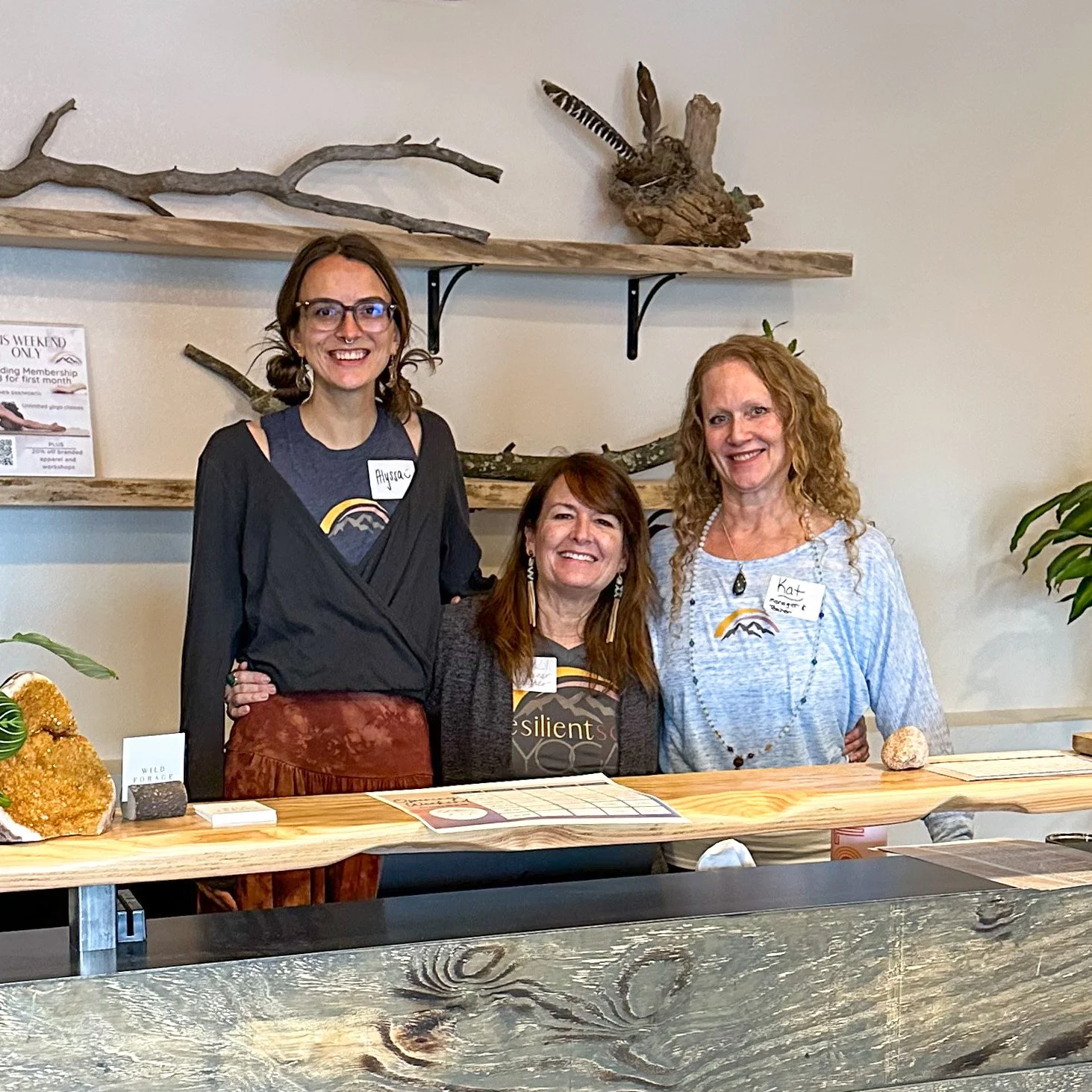 Three smiling women standing behind a wooden table at an indoor event, with decorative wood branches and feathers on the wall behind them.