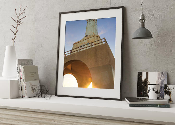 Framed photo of a lighthouse viewed from below, with sunlight shining through the arch at its base, on a white shelf with decor and books.