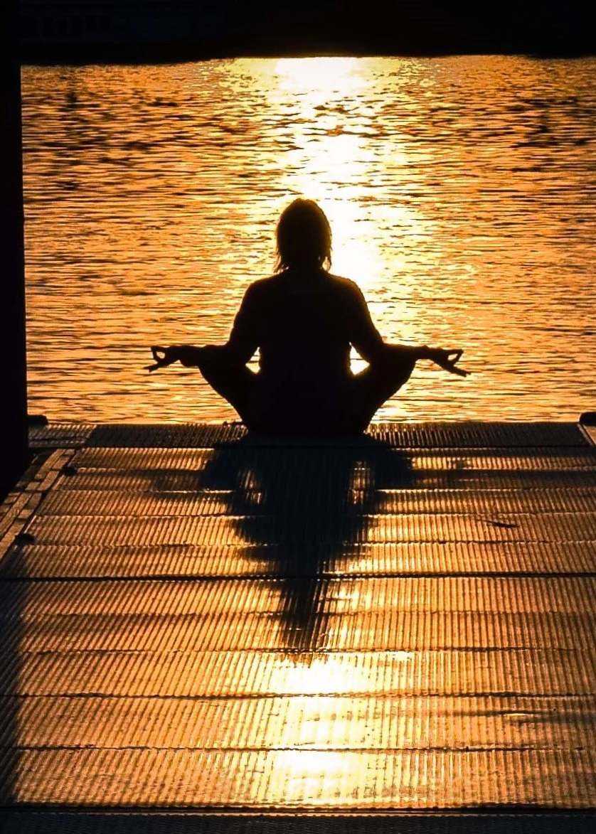 Silhouette of a person practicing yoga on a dock at sunset, facing a calm body of water.