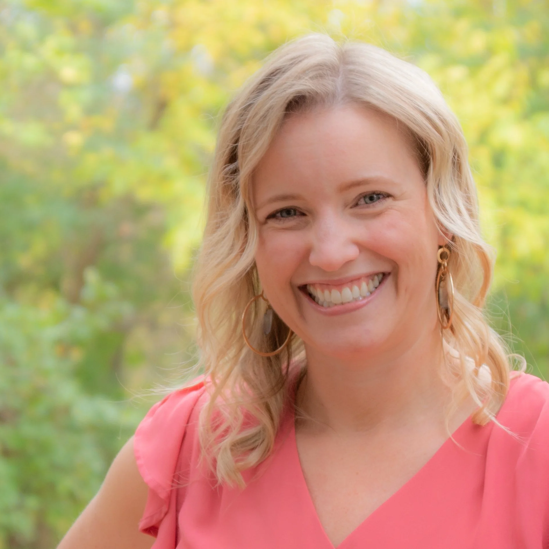 Close-up of a smiling woman with blond hair wearing a pink top, outdoors in a green, leafy background.