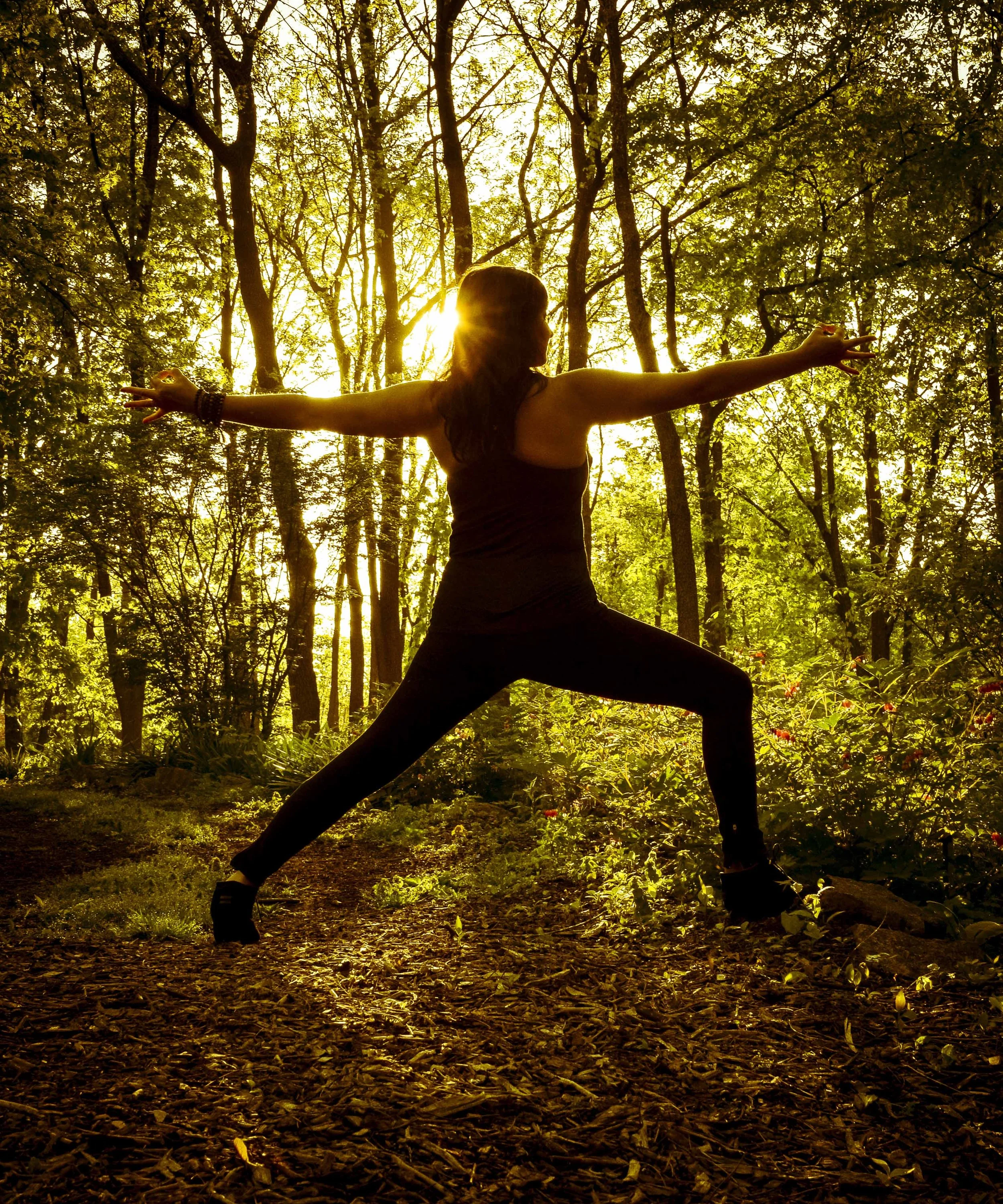A woman doing yoga outdoors in a forest with sunlight shining through trees.