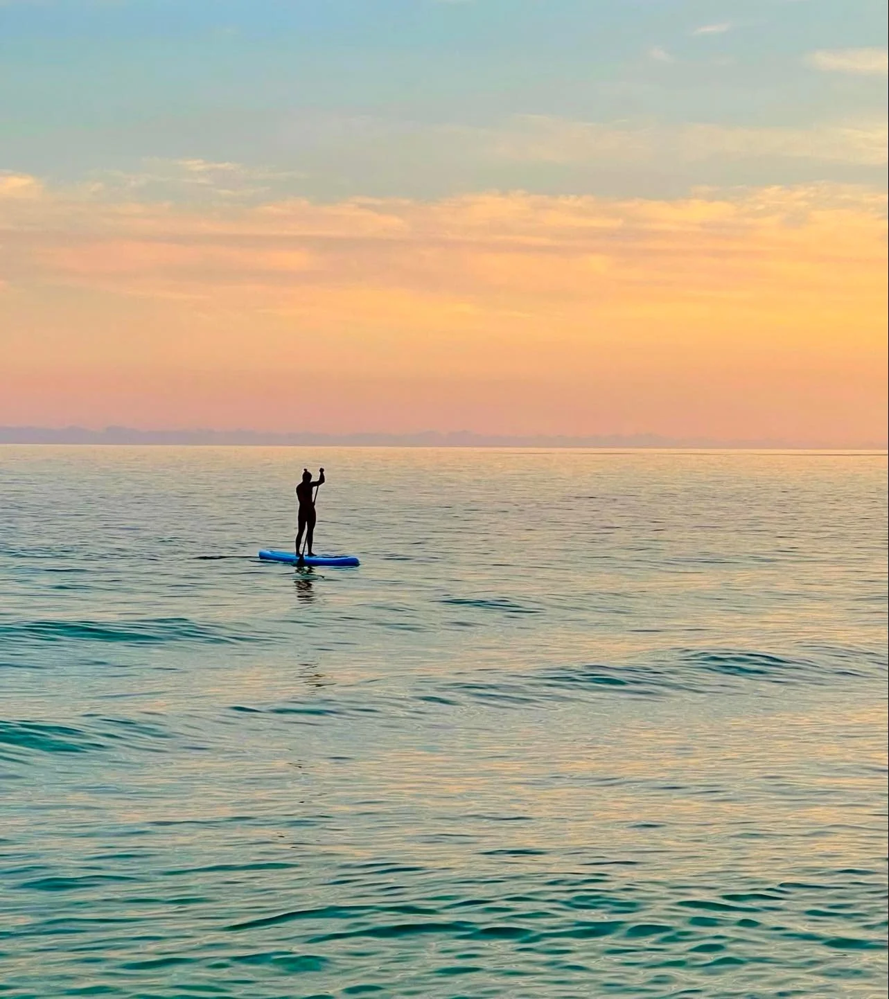 A person standing on a paddleboard in the water during sunset, holding a paddle.