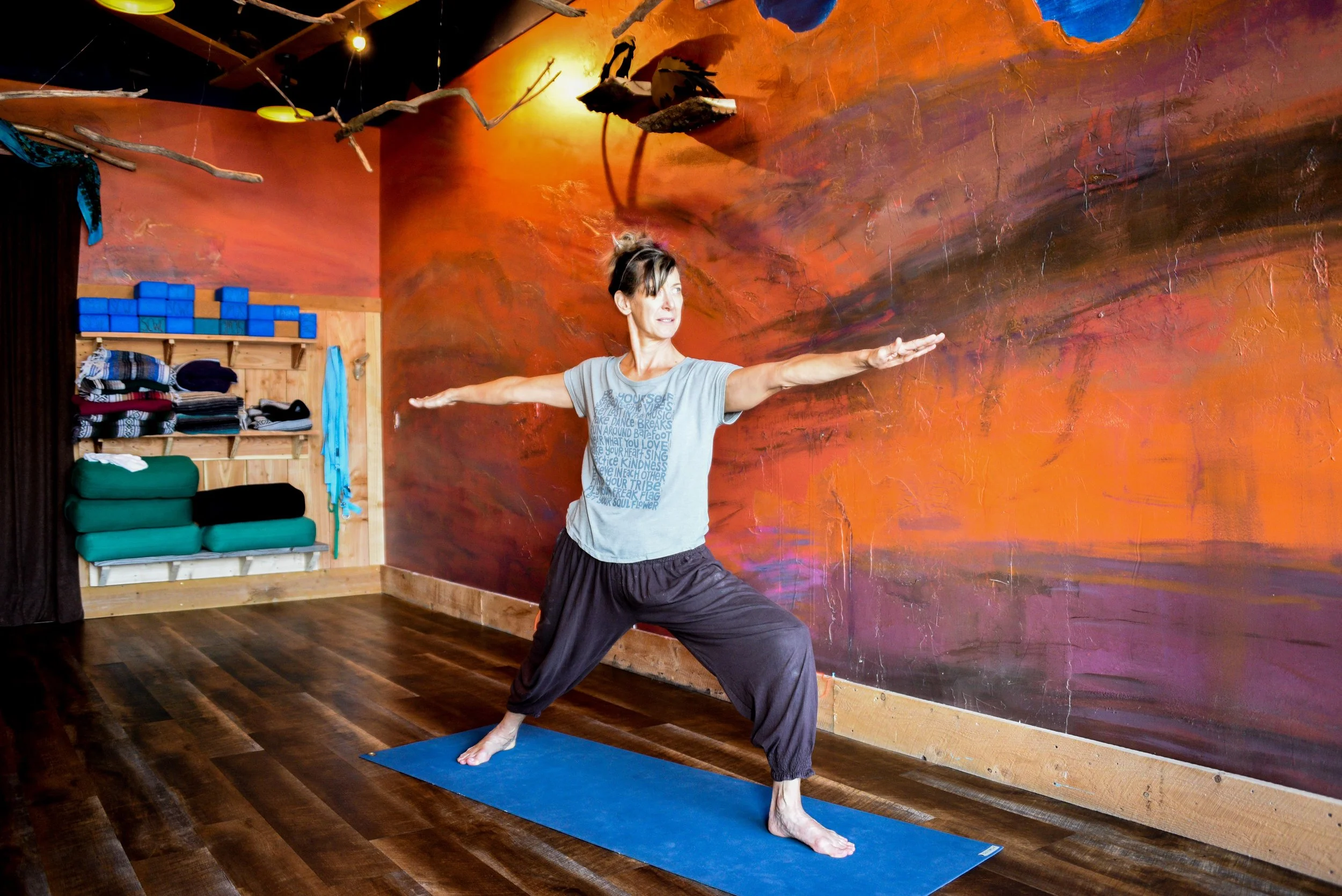 A woman practicing yoga indoors on a blue yoga mat, standing in a side warrior pose with arms extended, in a room with a colorful sunset mural on the wall and shelves of yoga props.