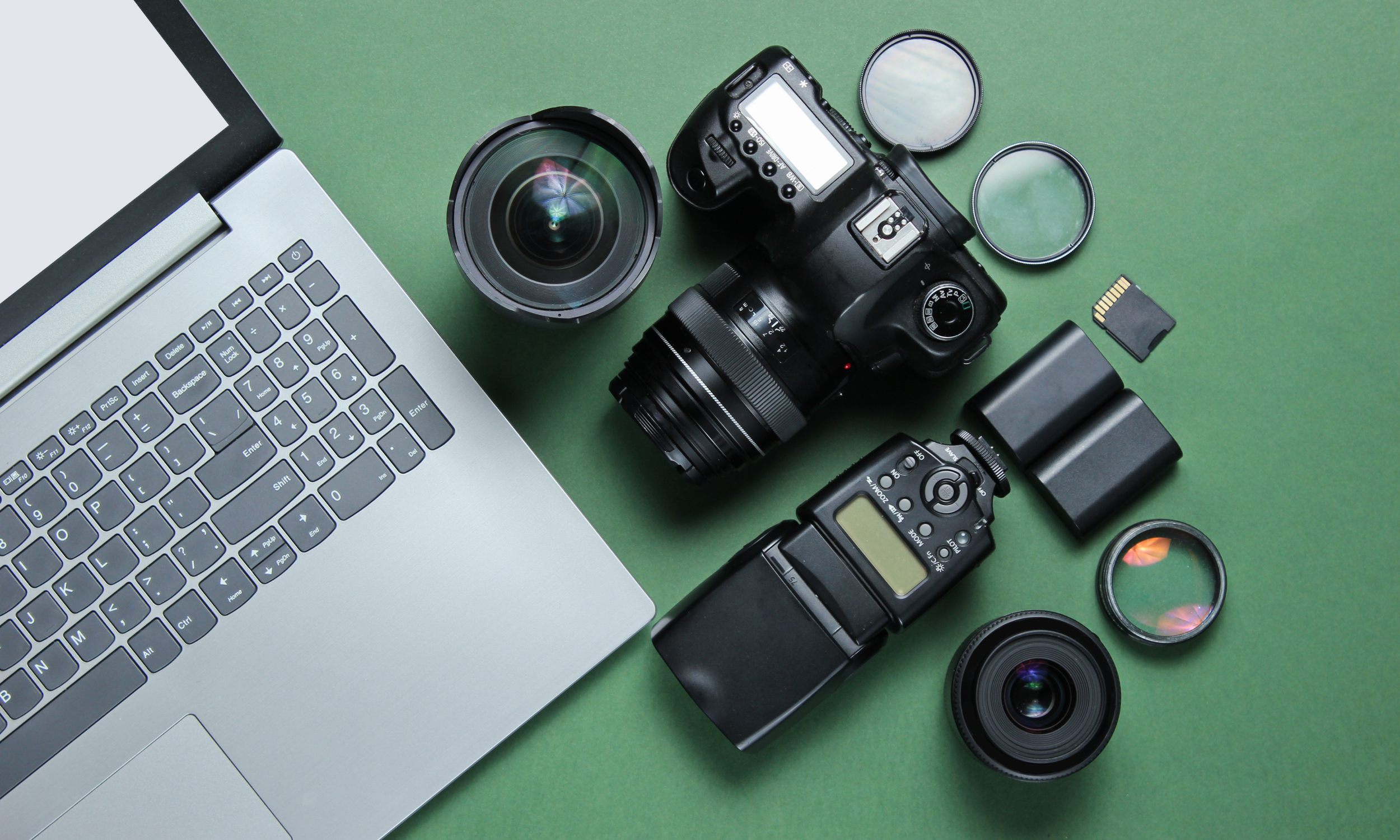 A laptop, two camera lenses, a digital camera, camera filters, a memory card, and camera batteries on a green surface.
