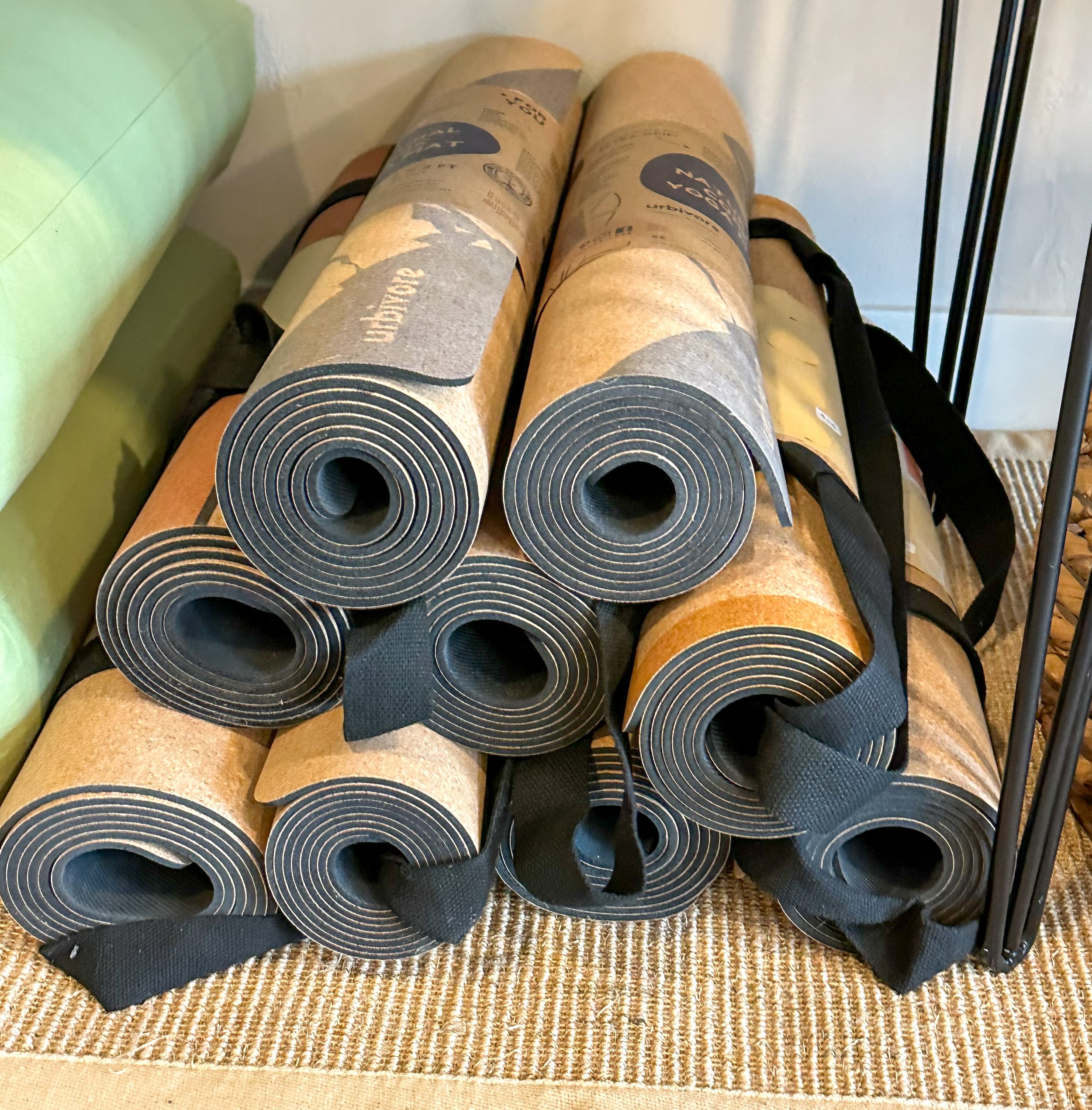 Multiple rolled-up yoga mats stacked on a woven mat, with a green cushion and a black metal frame nearby.