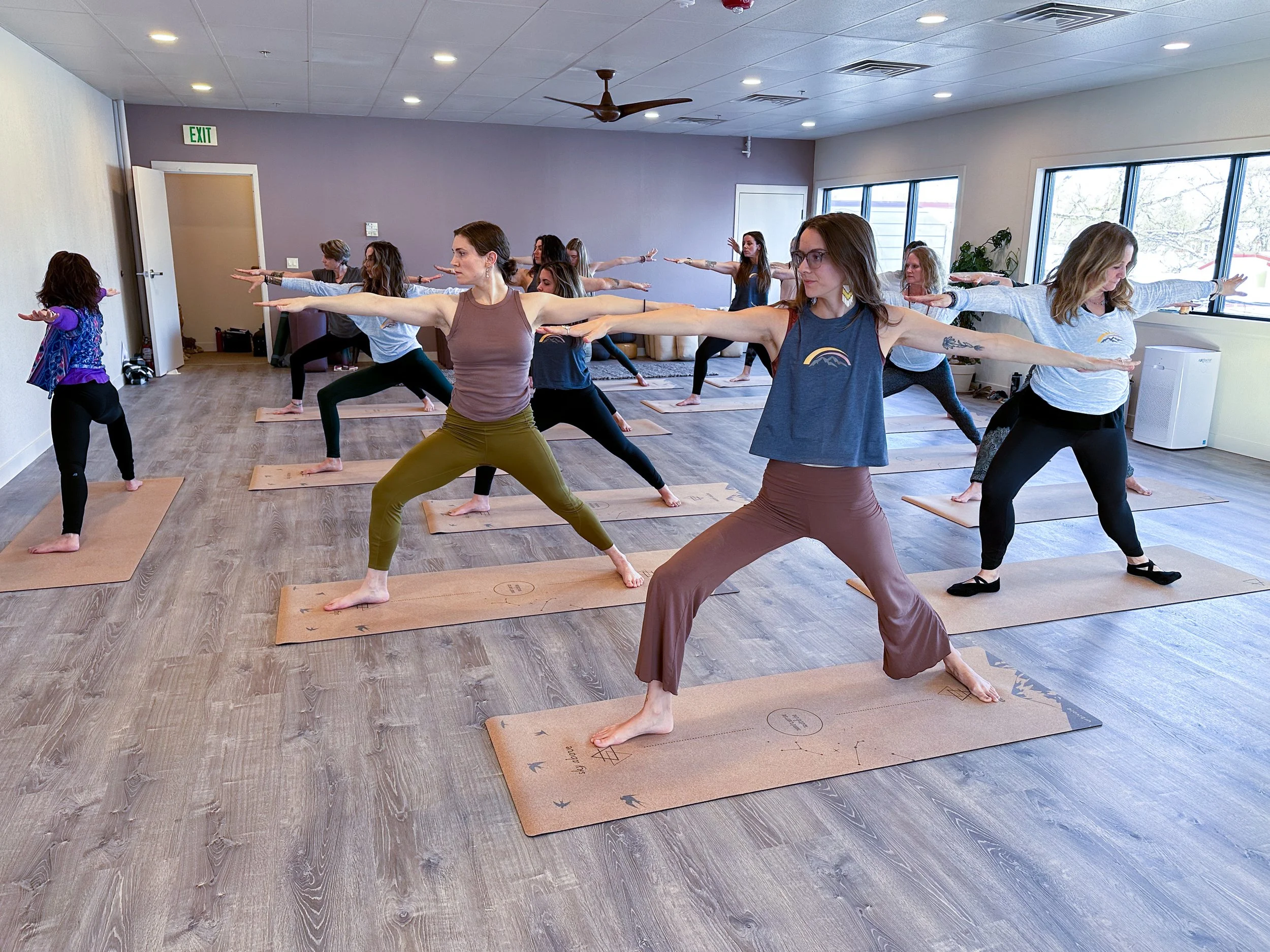 A group of women participating in a yoga class in a studio, performing Warrior Pose with arms extended, on mats.