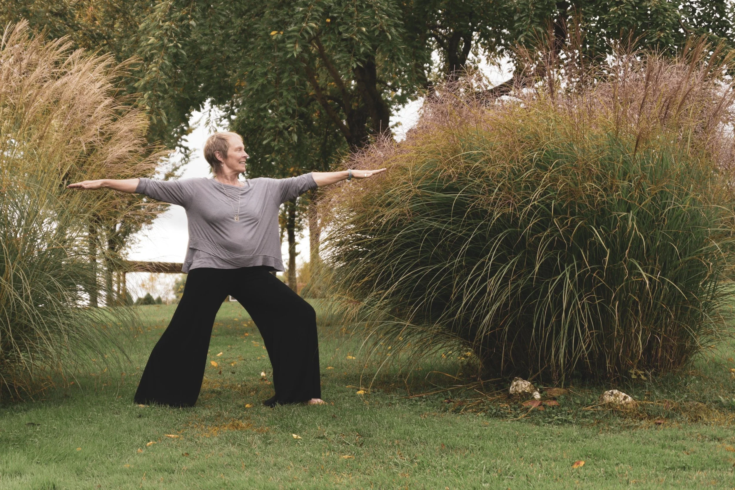 A woman practicing Warrior II yoga pose outdoors, standing with arms stretched out and smiling, surrounded by tall ornamental grasses and trees.