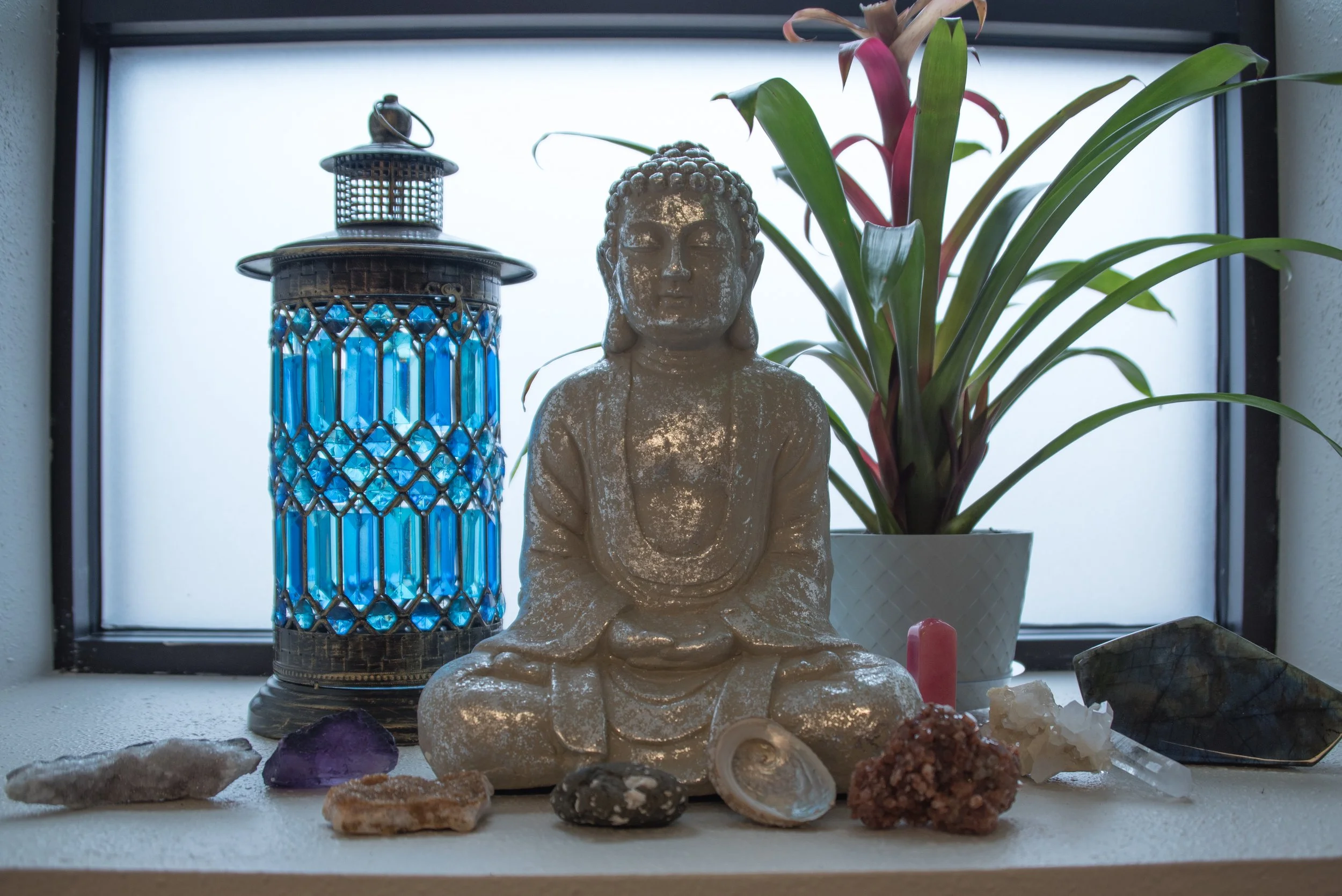 A small Buddha statue sitting on a shelf with various crystals and stones around it, a potted plant with green and red leaves, and a blue lantern, all set against a frosted window in the background.