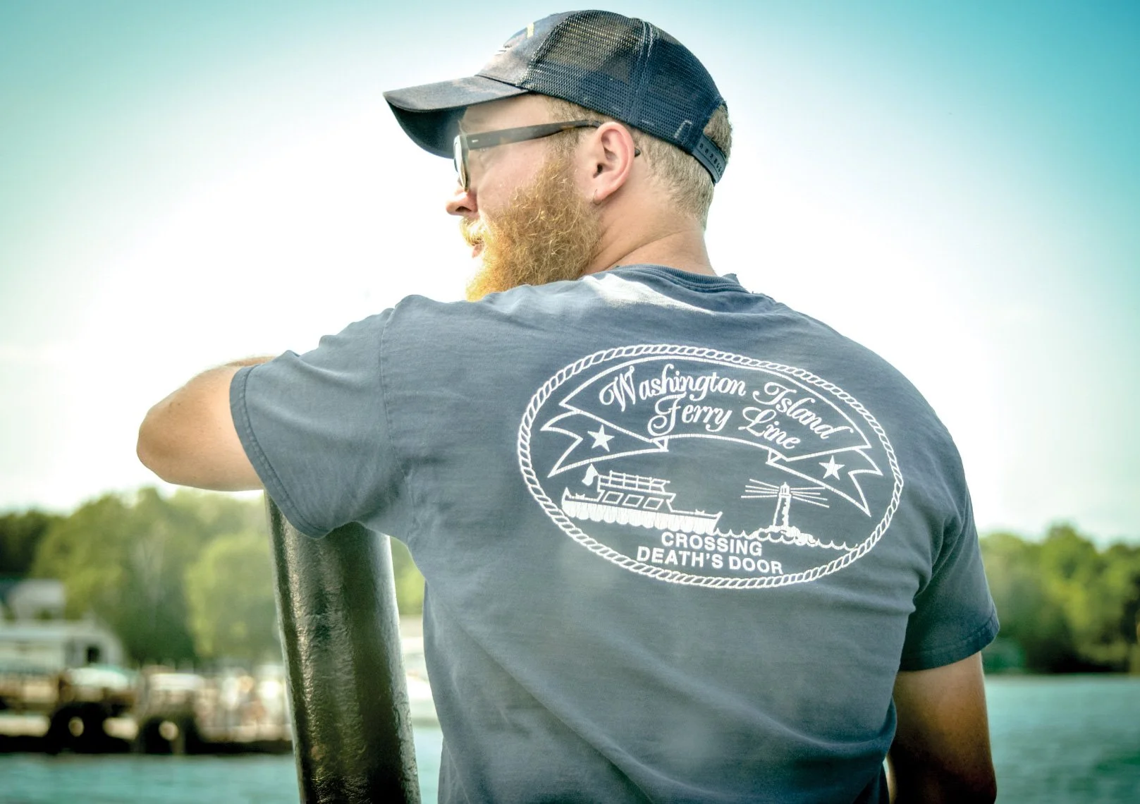 Man with a beard wearing a gray t-shirt with a Washington Island Ferry Line graphic and a cap, standing near water with trees in the background.