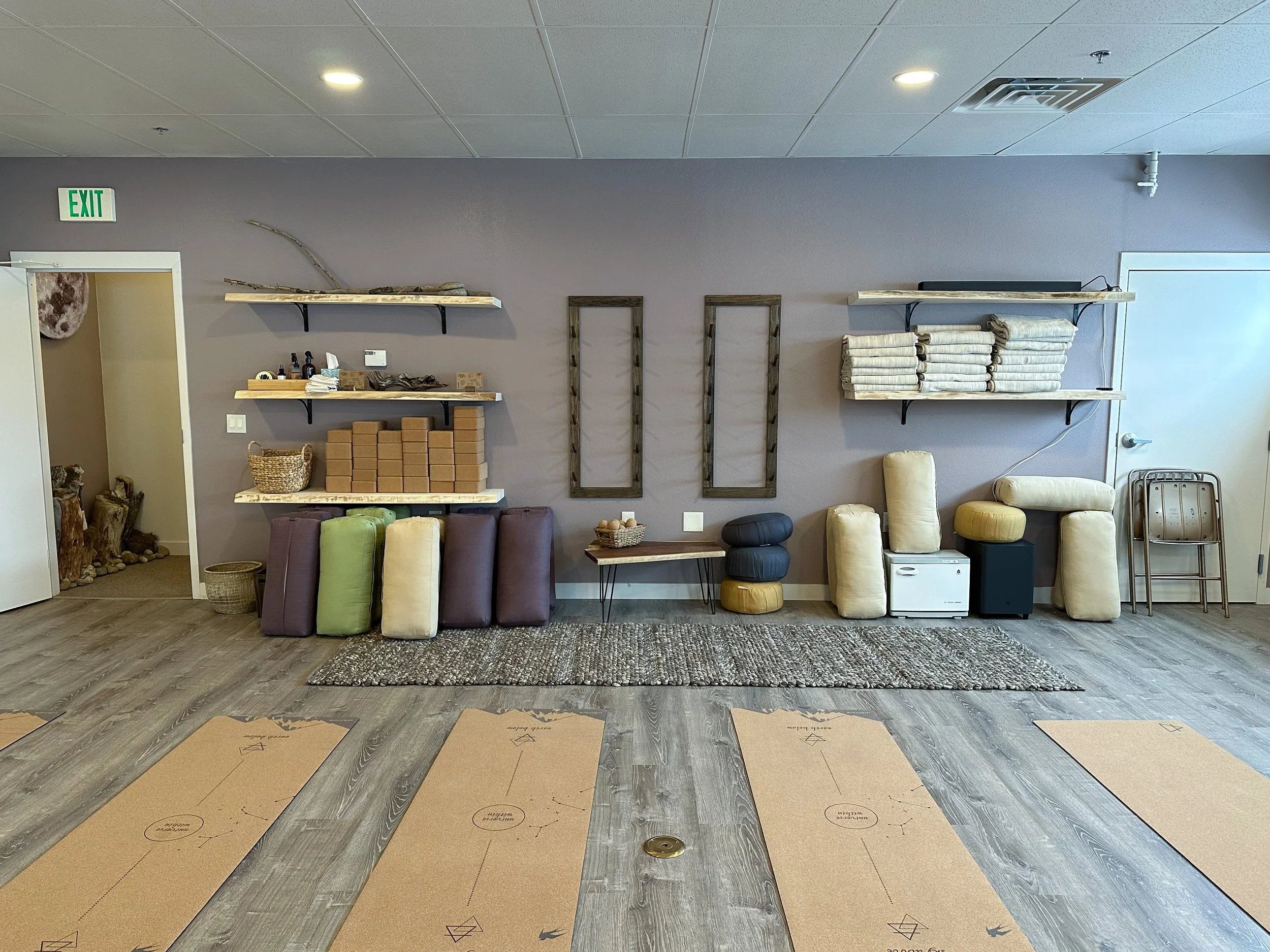 Yoga mats laid out on a wooden floor in a yoga studio, with various shelving and storage units on the wall holding towels, blocks, and decorative items.