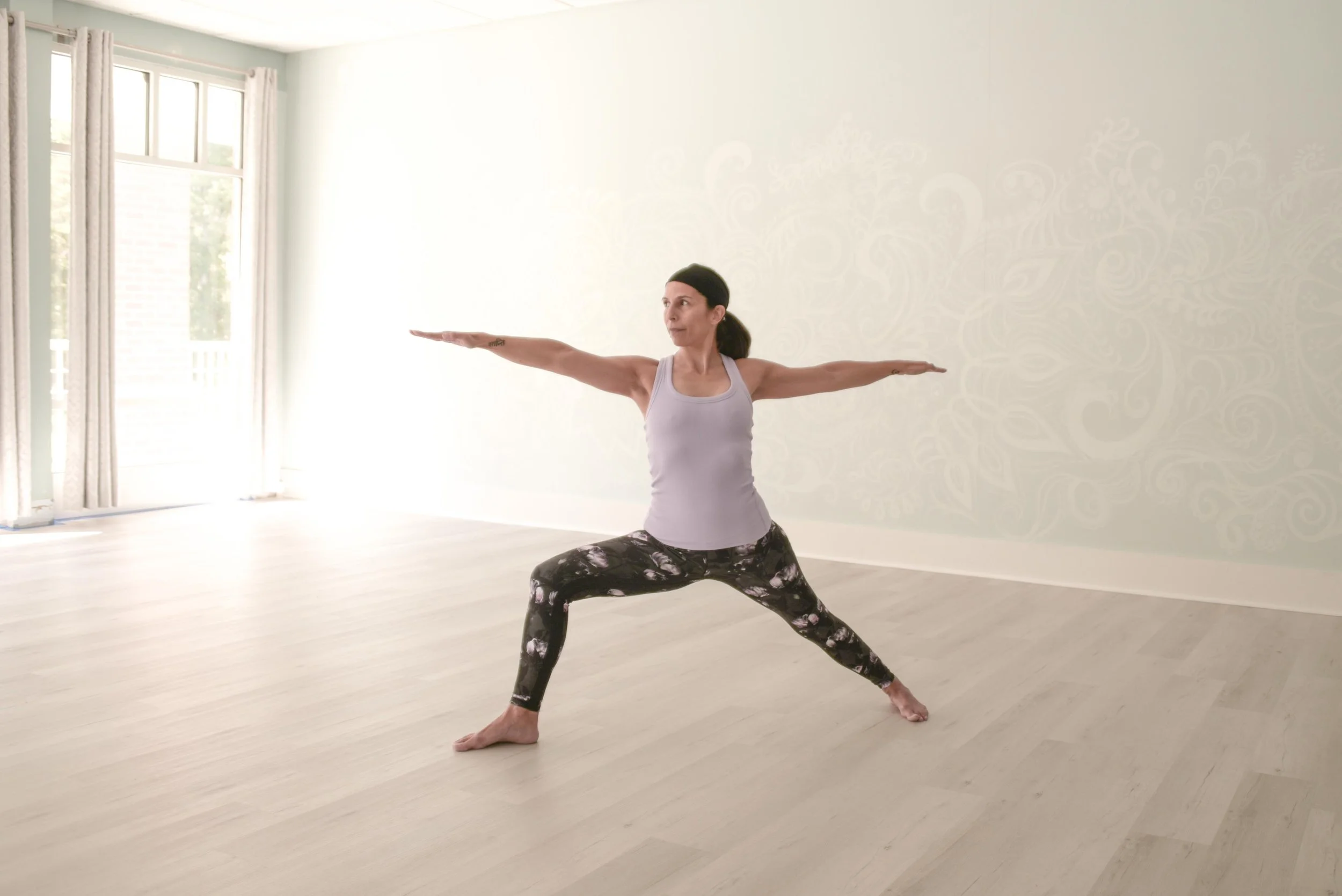 Woman practicing Warrior II yoga pose in a bright room with light-colored walls and wooden flooring, with her arms extended and legs apart in a warrior pose.