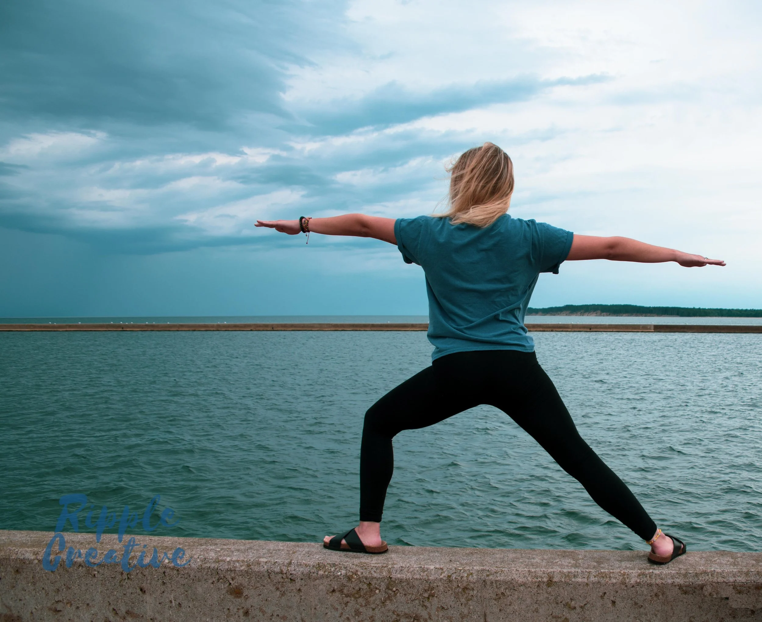 Woman doing yoga pose on a concrete pier near the water, with a cloudy sky overhead.