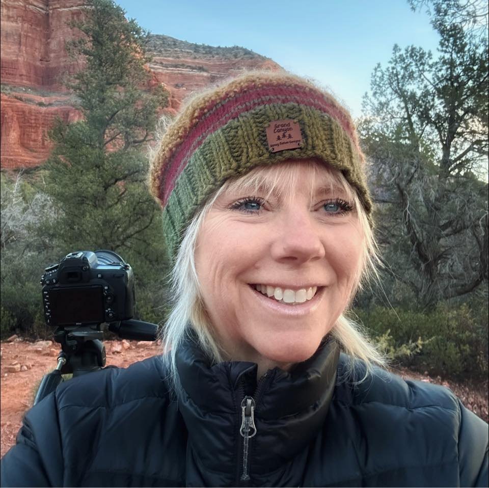 A smiling woman wearing a multicolored beanie and a black jacket in a desert landscape with red rock cliffs and trees.