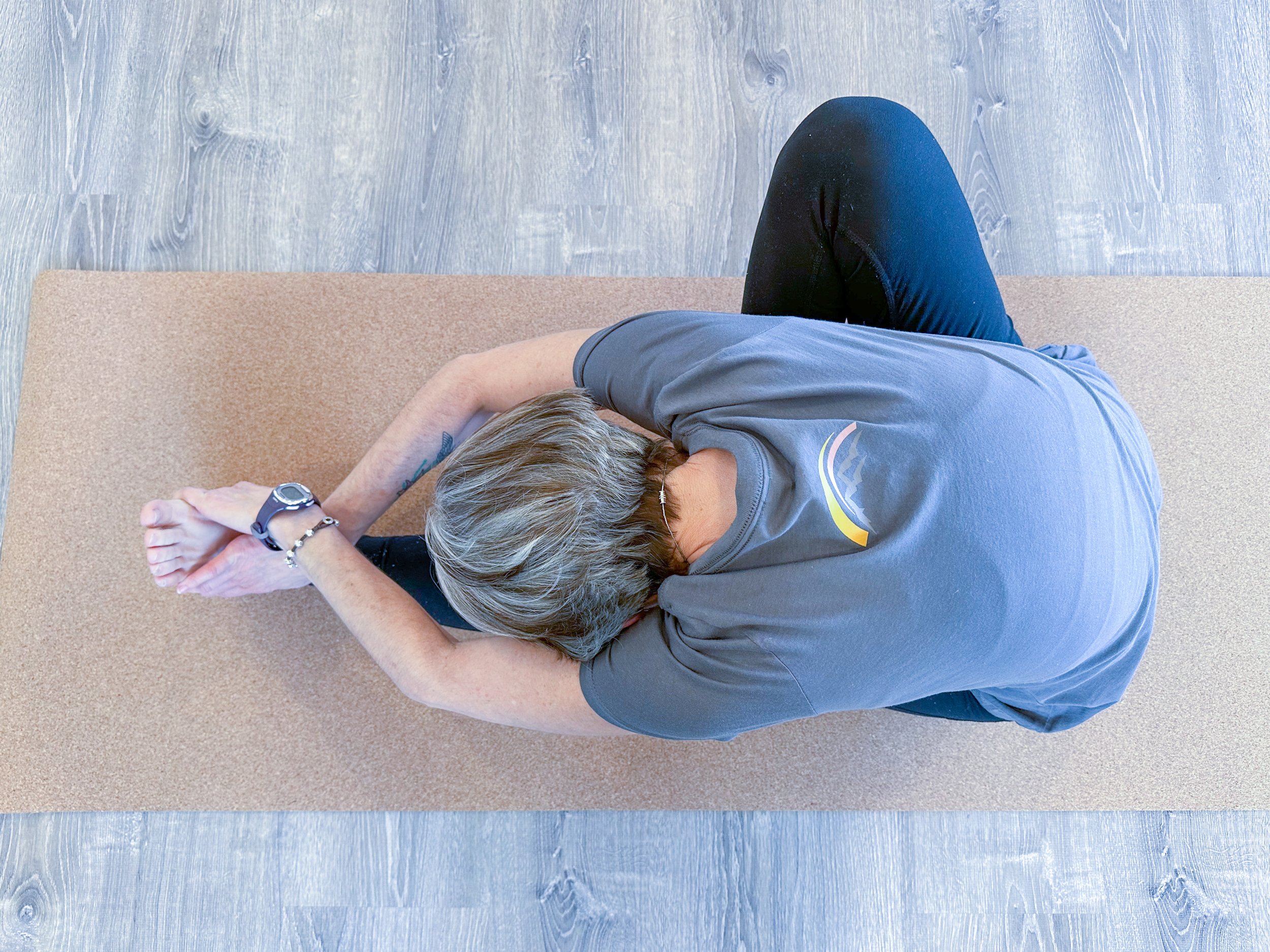 A person with short gray hair practicing yoga on a beige mat, sitting cross-legged with arms stretched overhead, grasping their left wrist with right hand, in a yoga pose.