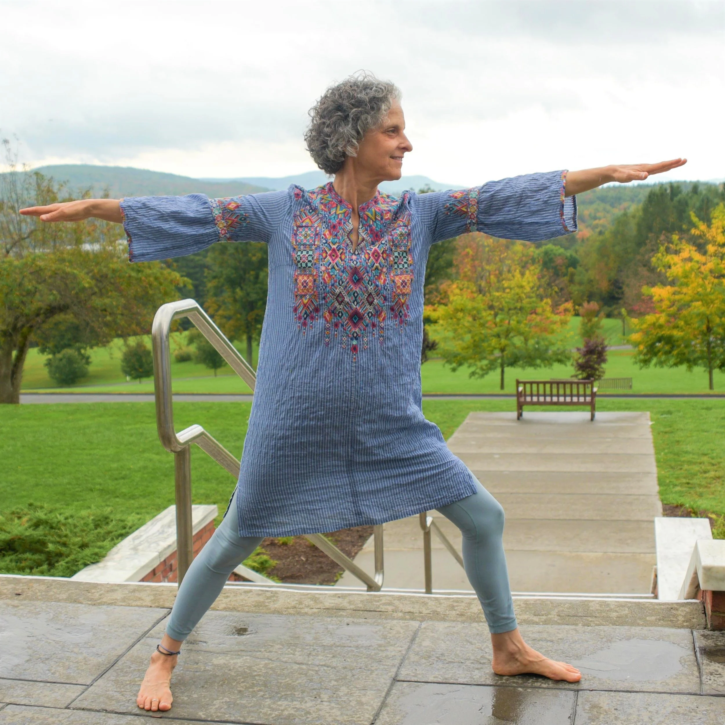 Older woman in colorful embroidered tunic practicing yoga outdoors on stone stairs in park with trees and mountains in background.