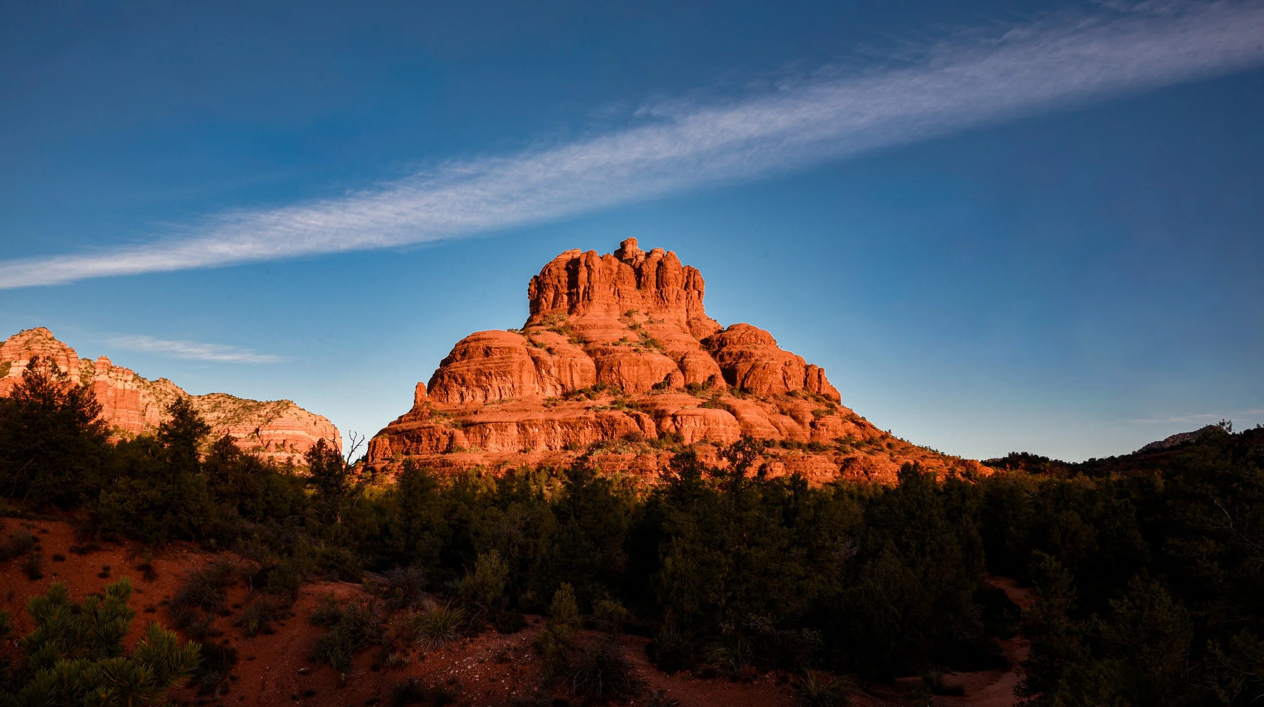 Red rock formation in a desert landscape with green trees and a clear blue sky.