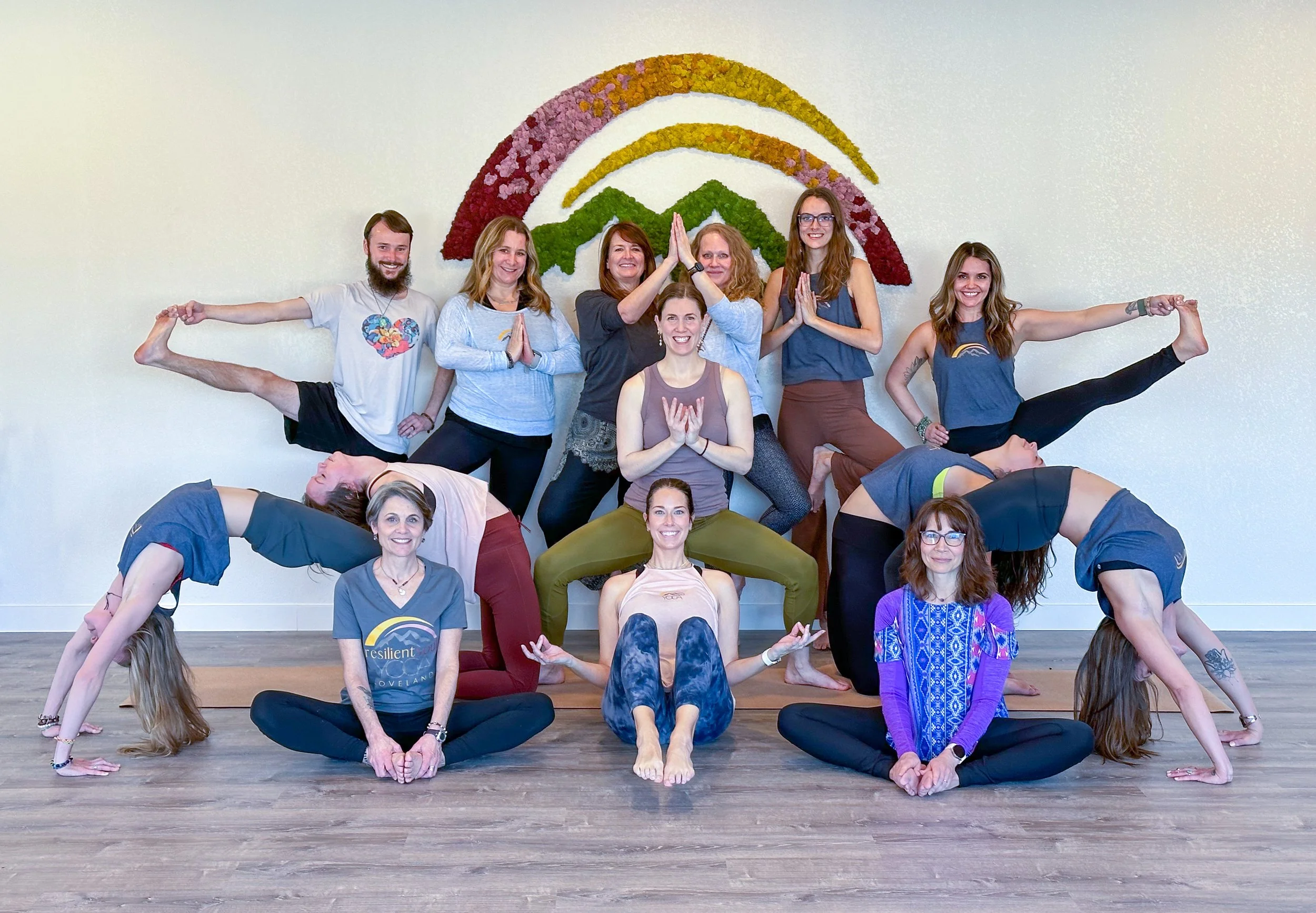 Group of people practicing yoga indoors, forming a human pyramid in front of a wall with a colorful flower and rainbow decoration.