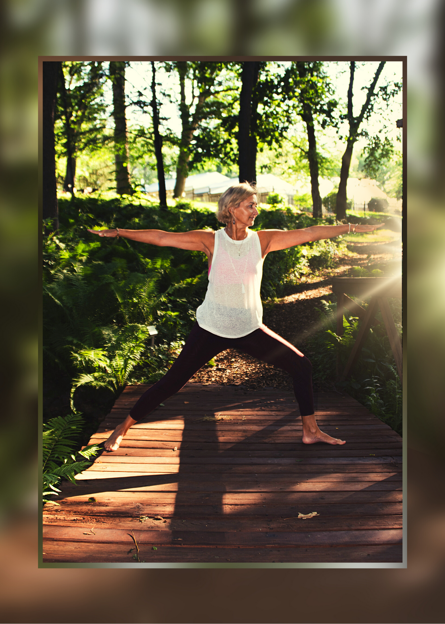 An older woman practicing yoga outdoors on a wooden deck surrounded by trees and greenery, performing a warrior II pose with arms outstretched.